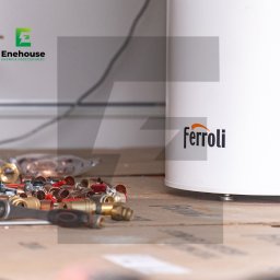 Enehouse Rafał Sołek - Biały zbiornik Ferroli z osprzętem hydraulicznym, złączkami miedzianymi i narzędziami na podłodze. Widoczny kabel zasilający i logo Enehouse.