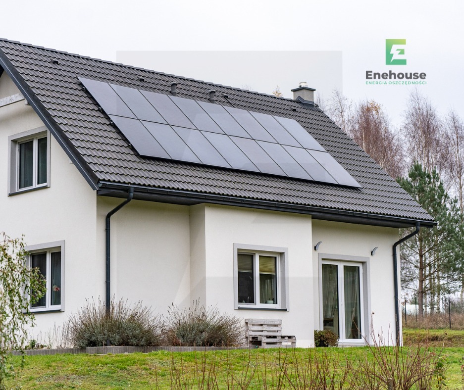 Biały dom jednorodzinny z ciemną dachówką i panelami fotowoltaicznymi na dachu. Widok z poziomu gruntu, pochmurny dzień. Logo firmy w prawym górnym rogu.