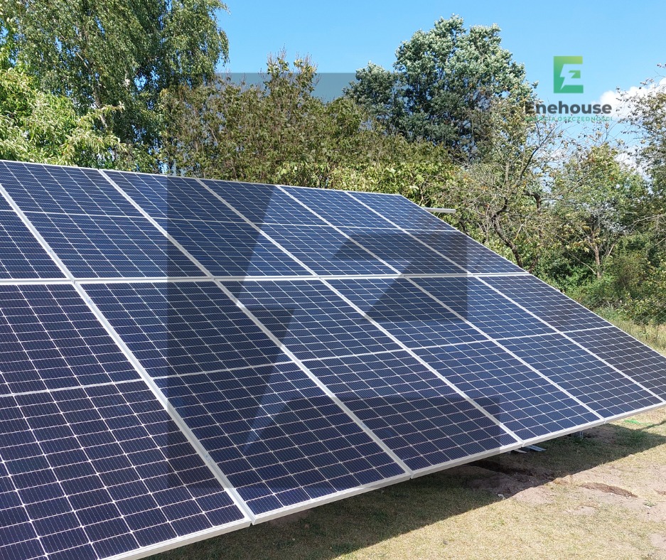 Instalacja fotowoltaiczna na gruncie w słoneczny dzień. Widoczne panele w układzie rzędowym, z logo firmy w prawym górnym rogu. Tło stanowią drzewa i błękitne niebo.