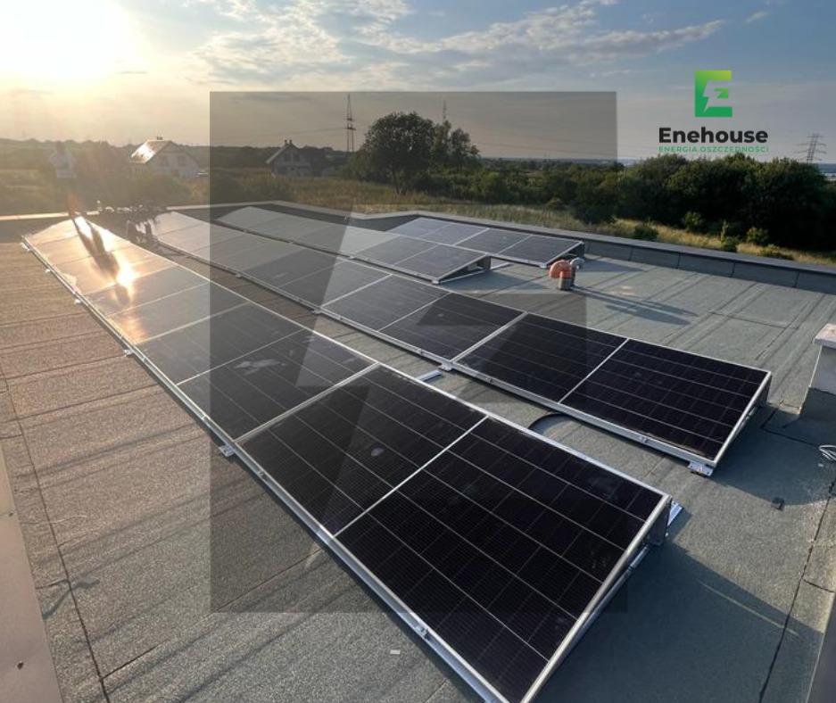 Instalacja fotowoltaiczna na płaskim dachu, widoczne panele w rzędach, w tle budynki mieszkalne i zachodzące słońce. Widoczny cień osoby montującej panele.