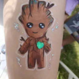 Rysunek na udzie dziecka przedstawia postać Groot z filmu Strażnicy Galaktyki, ozdobioną brokatem i zielonym sercem. Realizacja usługi malowania twarzy.