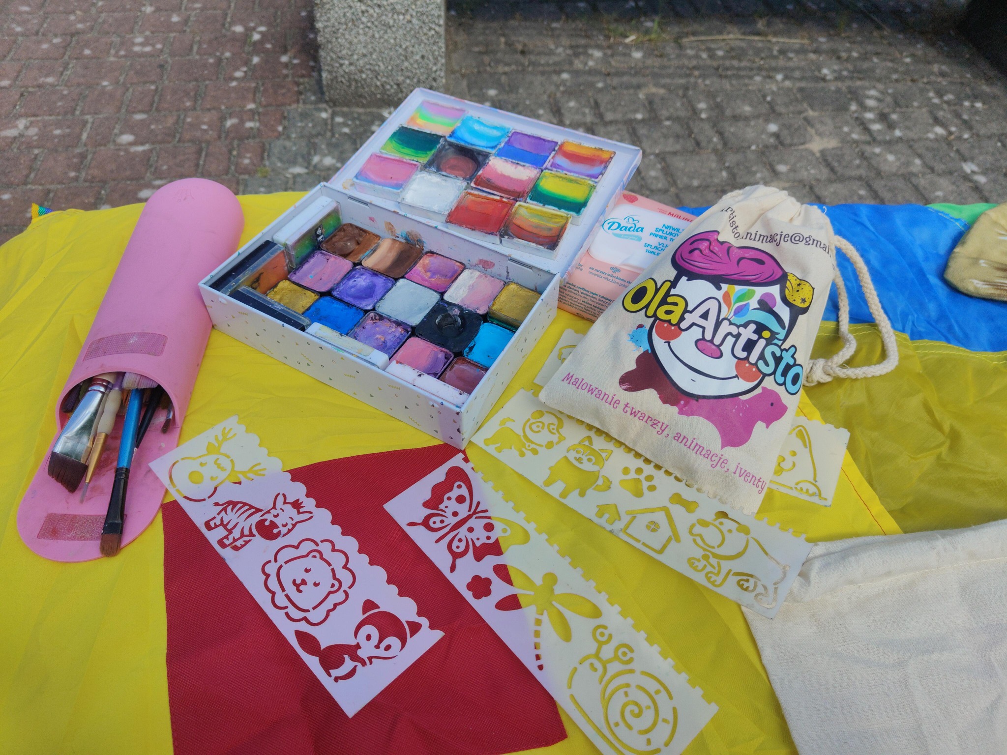 Zestaw do malowania twarzy: farby w kasetce, pędzle w etui, szablony ze zwierzętami i logo 'Ola Artisto' na worku, na kolorowym tle.