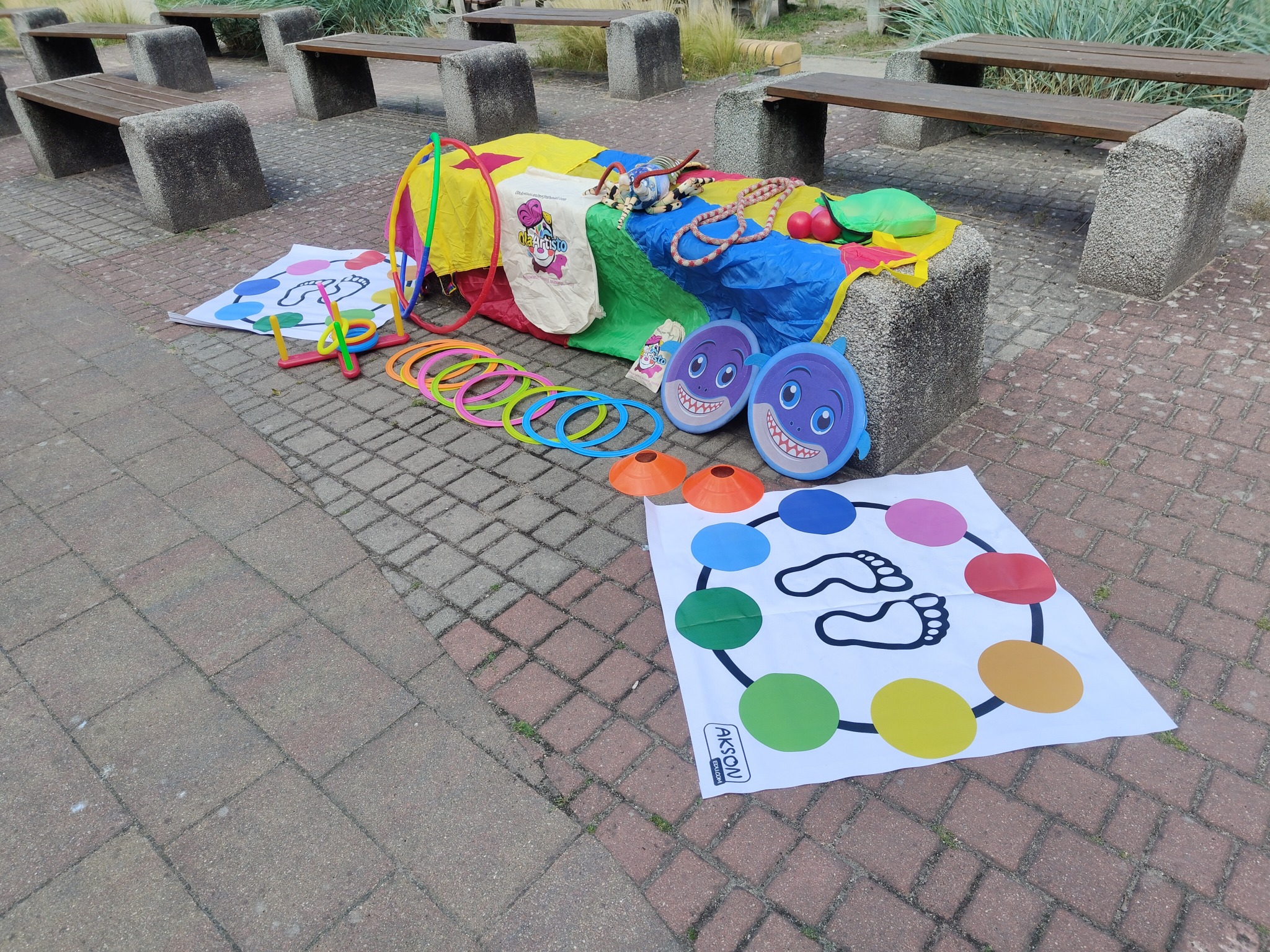 Kolorowy zestaw gier i akcesoriów dla dzieci na chodniku: tunel, koła, mata do twistera i rekwizyty, gotowe do animacji zabaw na świeżym powietrzu.