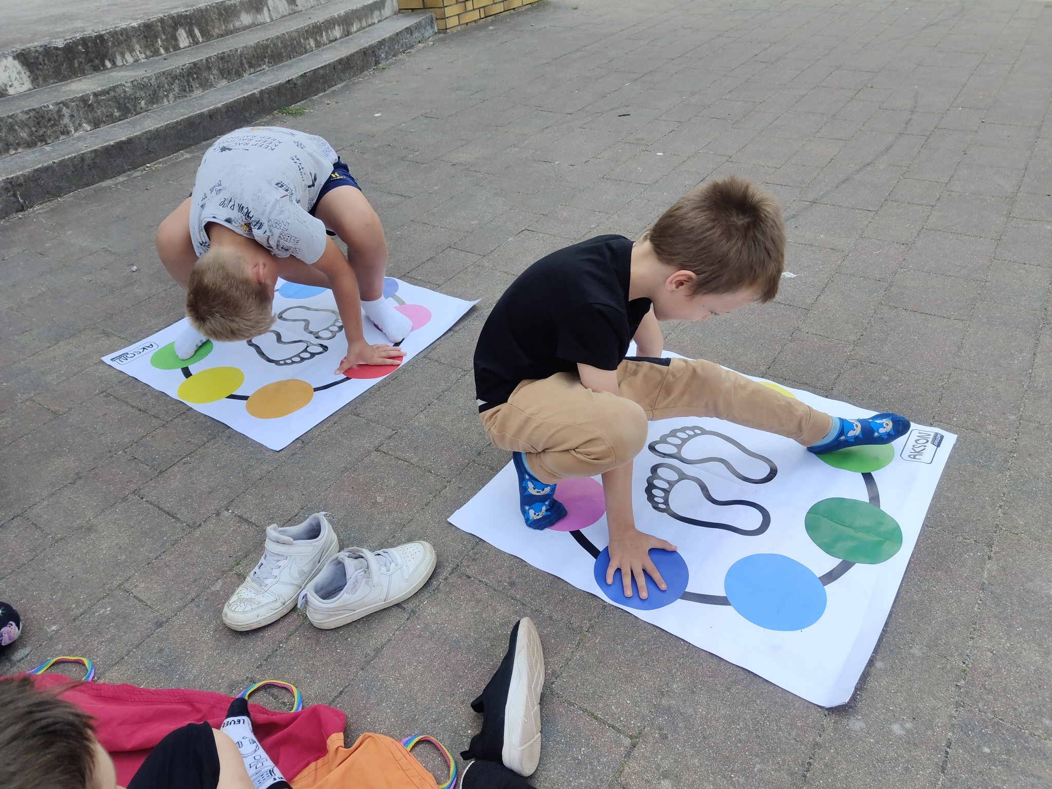 Dwóch chłopców gra w Twistera na chodniku, z butami i torbą obok. Jeden w szarej koszulce, drugi w czarnej, obaj skupieni na grze.