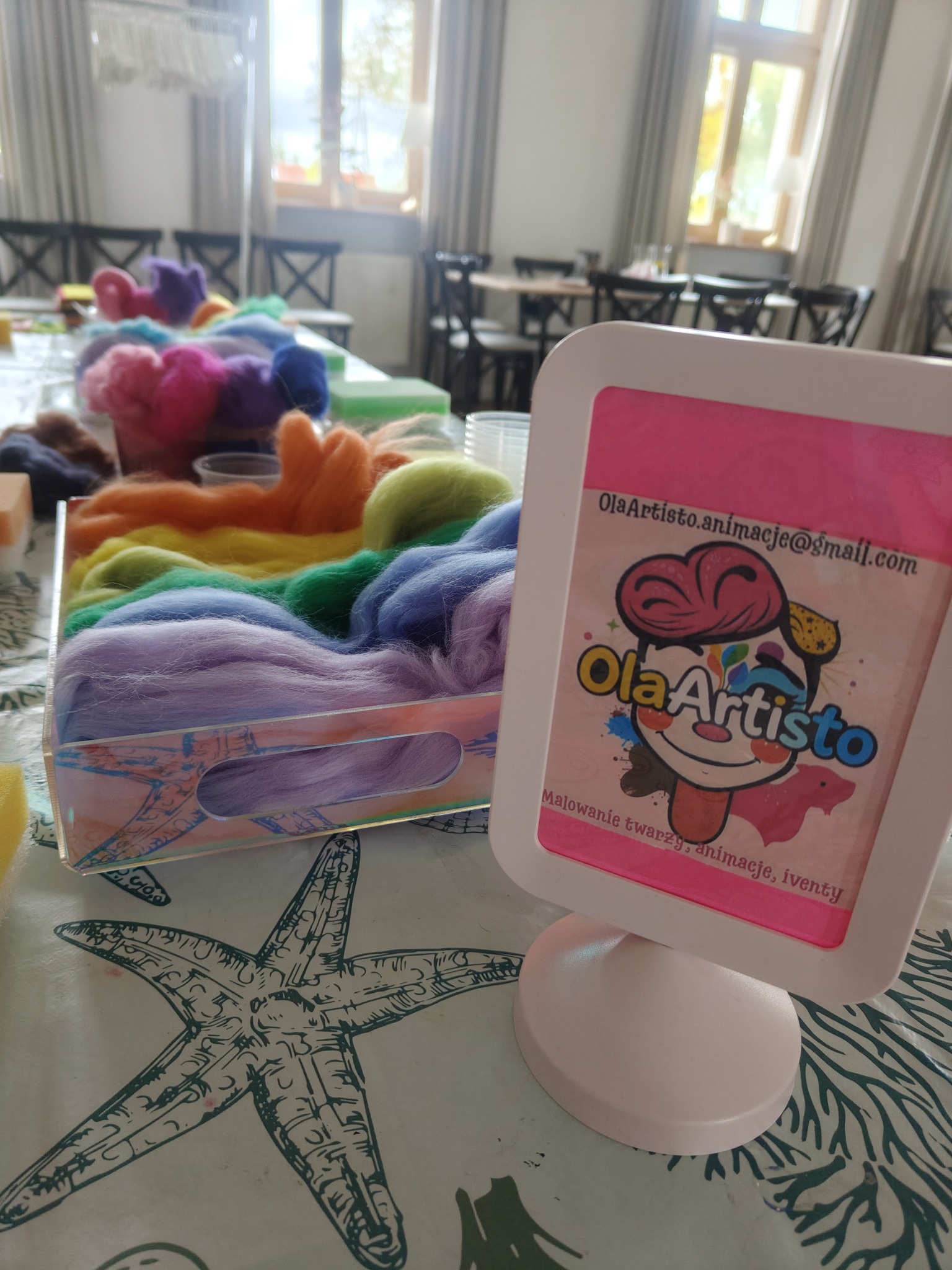 Kolorowa wełna w przezroczystym pojemniku obok ramki z logo 'Ola Artisto' i hasłem 'Malowanie twarzy, animacje, eventy' na stole z motywem rozgwiazd.