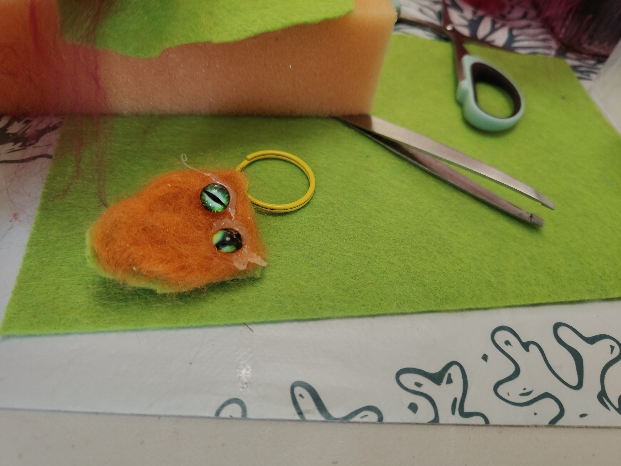 Ręcznie robiona pomarańczowa maskotka z filcu z zielonymi oczami na zielonej macie, obok pęseta i nożyczki. Etap tworzenia rekwizytu.