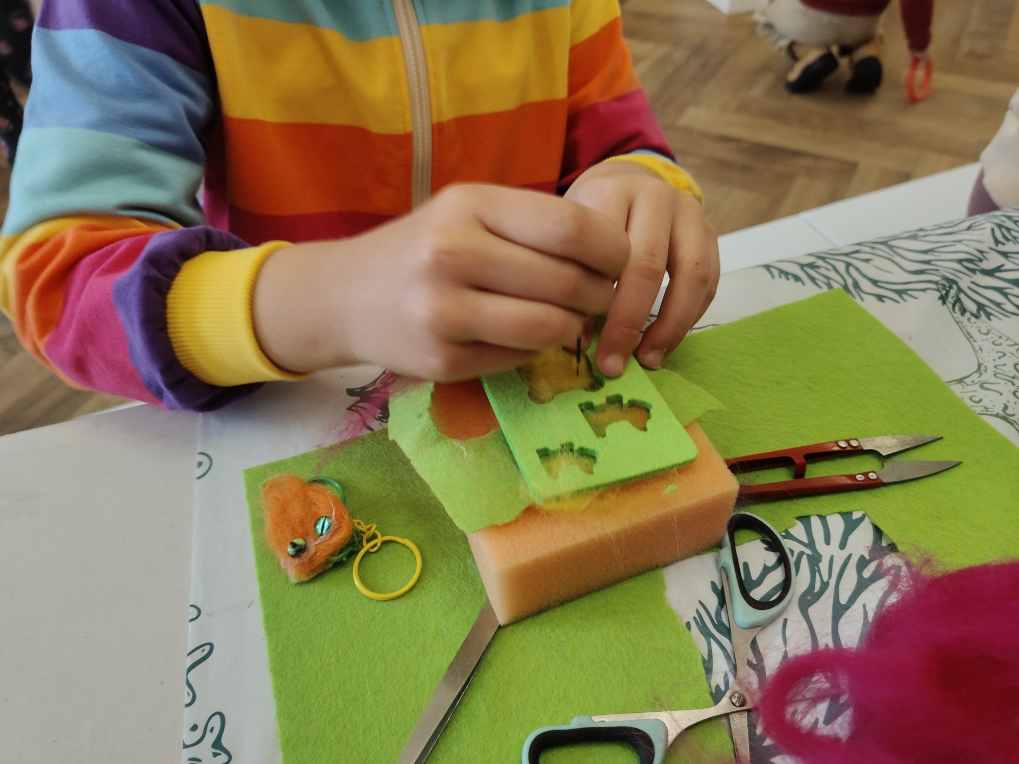 Dziecko szyje brelok z filcu na warsztatach rękodzielniczych. Widoczne materiały, narzędzia i gotowy breloczek w kształcie zwierzątka.