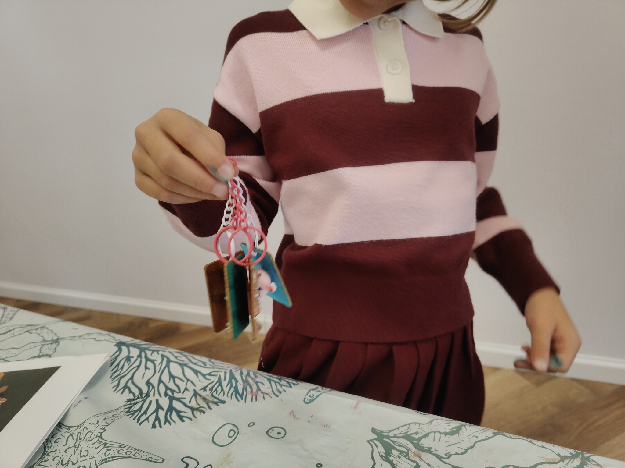 Dziecko trzyma breloczek z kartkami, prawdopodobnie wykonany podczas warsztatów plastycznych. Ubranie w paski różowo-bordowe. Blat stołu z motywem morskim.