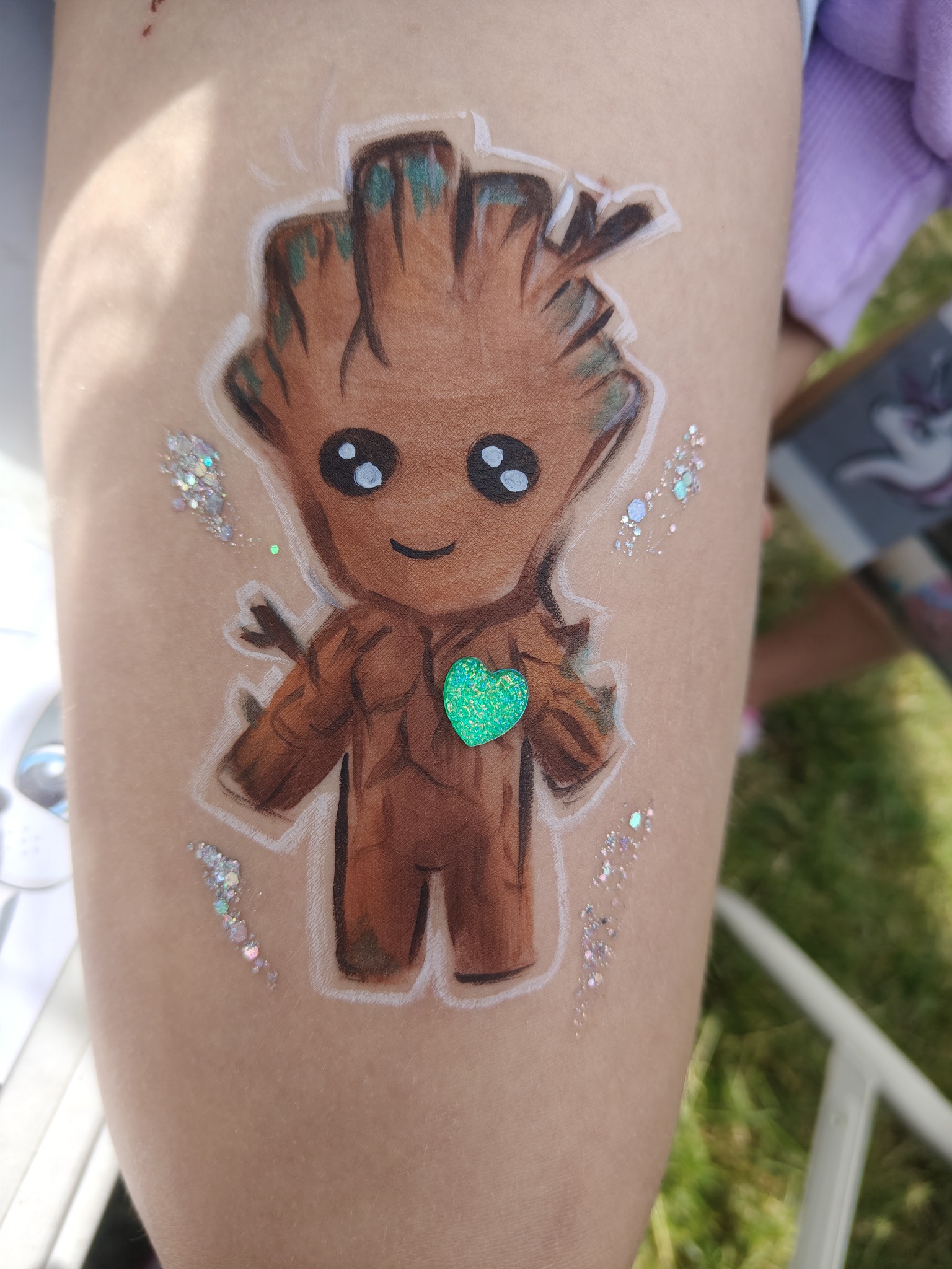 Rysunek na udzie dziecka przedstawia postać Groot z filmu Strażnicy Galaktyki, ozdobioną brokatem i zielonym sercem. Realizacja usługi malowania twarzy.