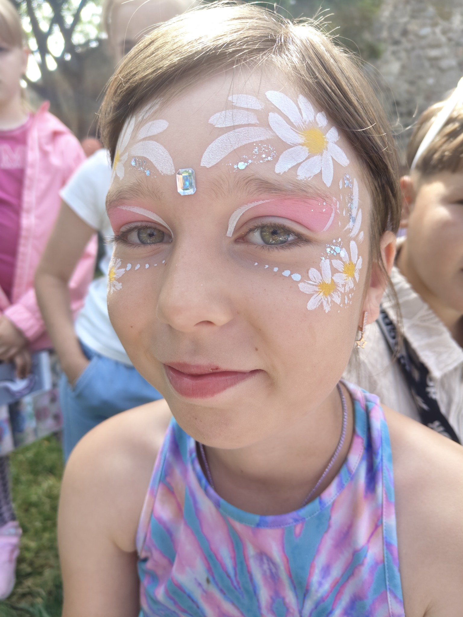 Uśmiechnięta dziewczynka z kwiatowym makijażem na twarzy i błyszczącym kamieniem na czole, otoczona innymi dziećmi w tle.