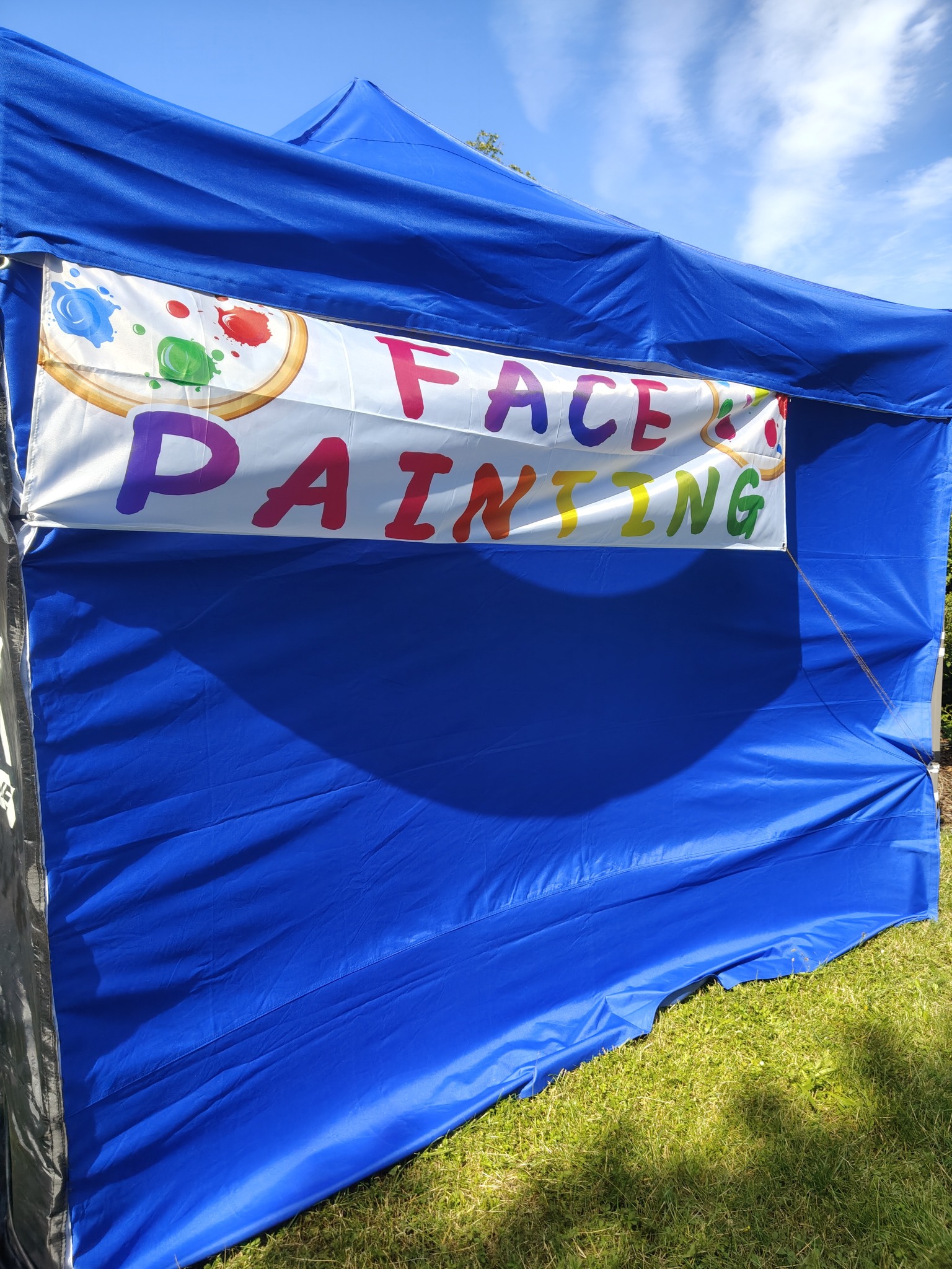 Niebieski namiot z banerem 'Face Painting' i paletą farb, ustawiony na trawie w słoneczny dzień. Kąt ujęcia od dołu, widoczne niebo.