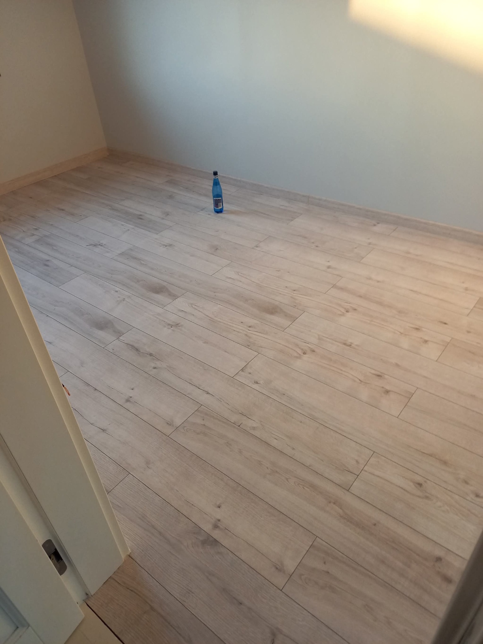 Jasne panele podłogowe ułożone w pokoju, widok z góry, butelka wody w tle. Fragment ościeżnicy po lewej stronie kadru.