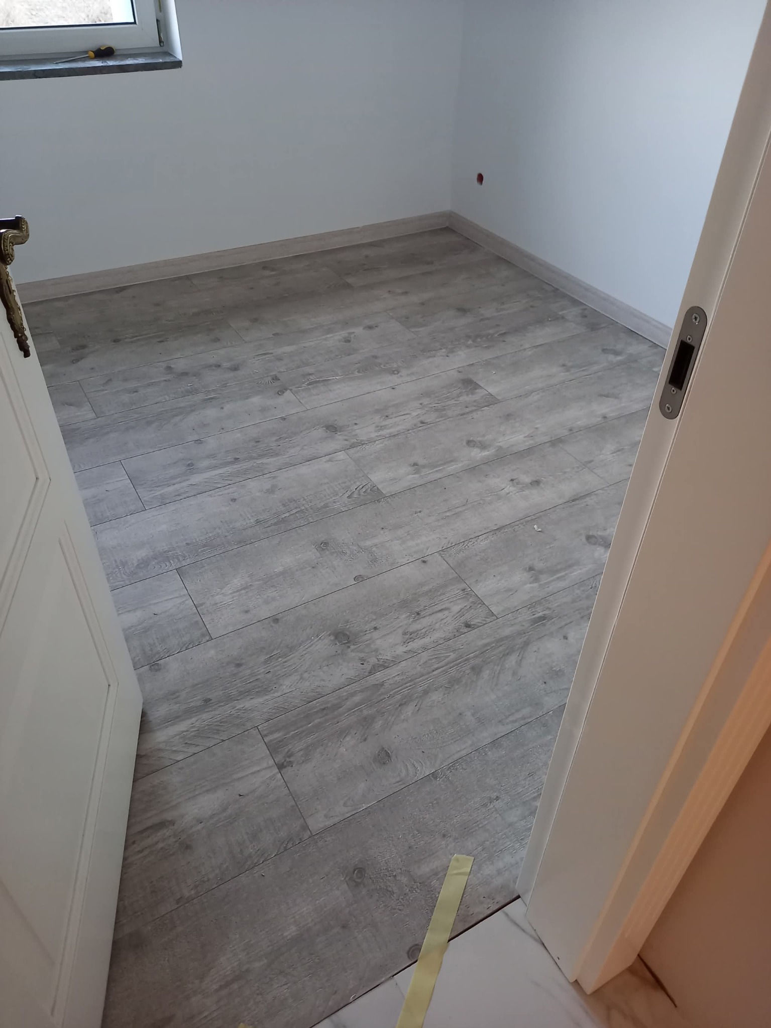 Jasne panele podłogowe imitujące drewno w nowym pokoju, widoczne z perspektywy otwartych drzwi. Świeżo pomalowane ściany i listwy przypodłogowe.