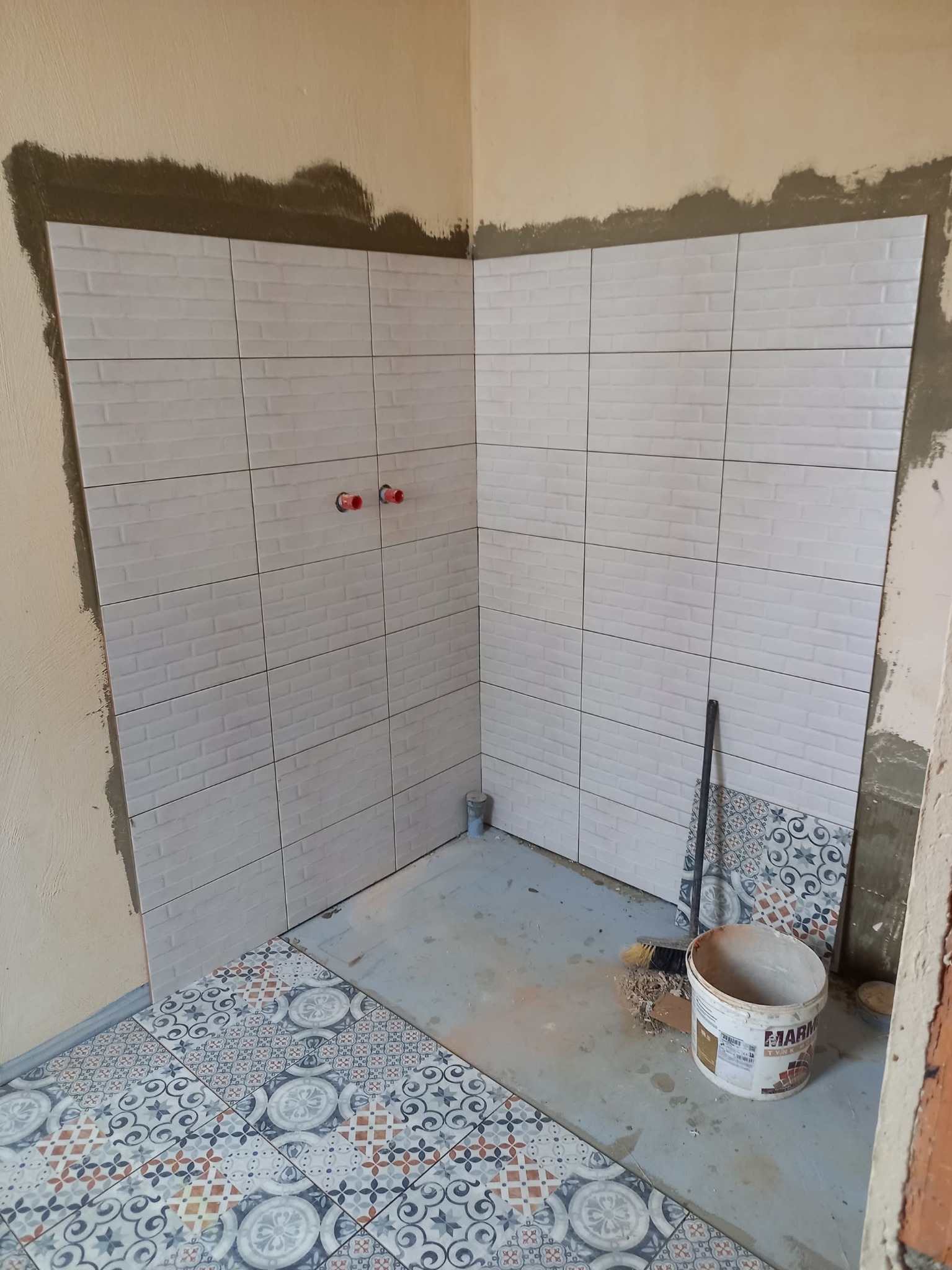 Fragment łazienki w trakcie remontu: białe, cegiełkowe płytki na ścianach, wzorzyste płytki podłogowe, widoczne narzędzia i wiadro. Etap prac wykończeniowych.