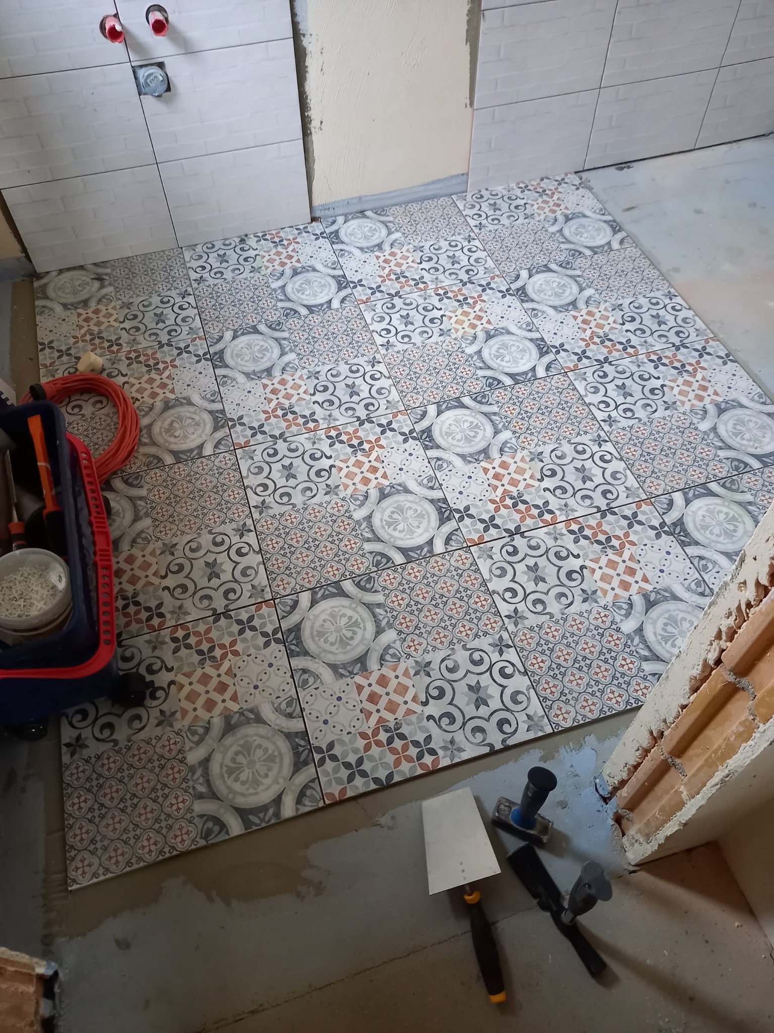 Ułożone płytki podłogowe z wzorem patchwork w łazience, widoczne narzędzia glazurnicze i fragment ściany z białymi płytkami. Prace w toku.