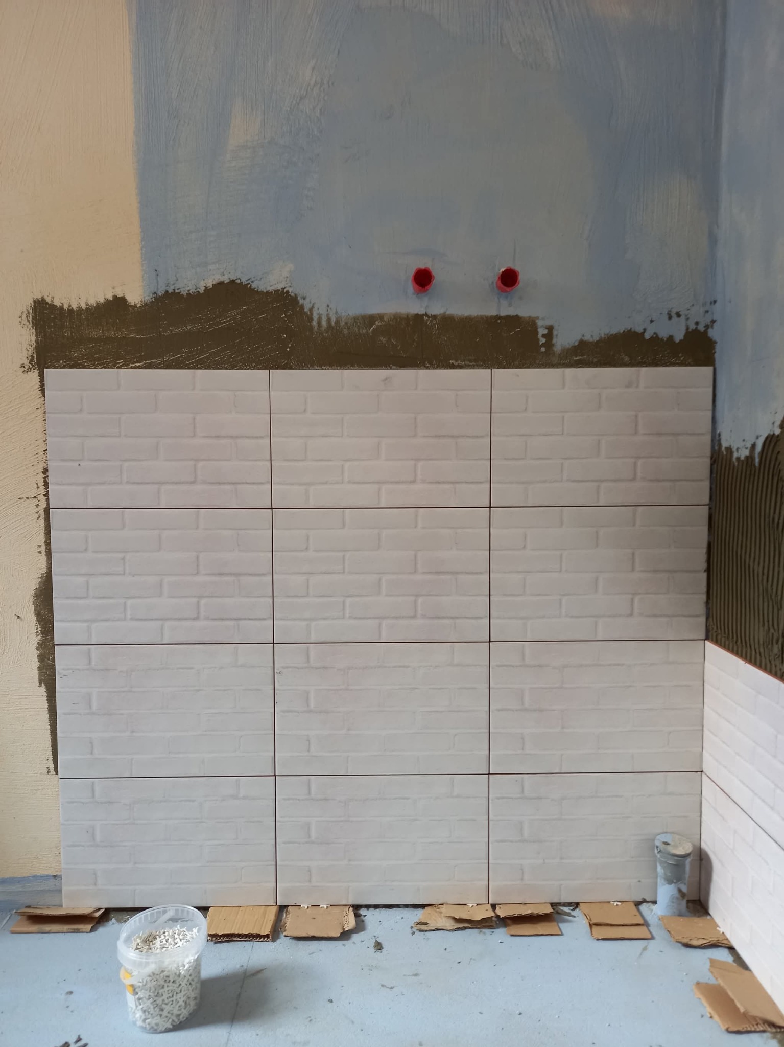 Ściana w trakcie układania białych, prostokątnych płytek imitujących cegłę. Widoczne czerwone końcówki rur, zaprawa i poziomowanie kartonami.