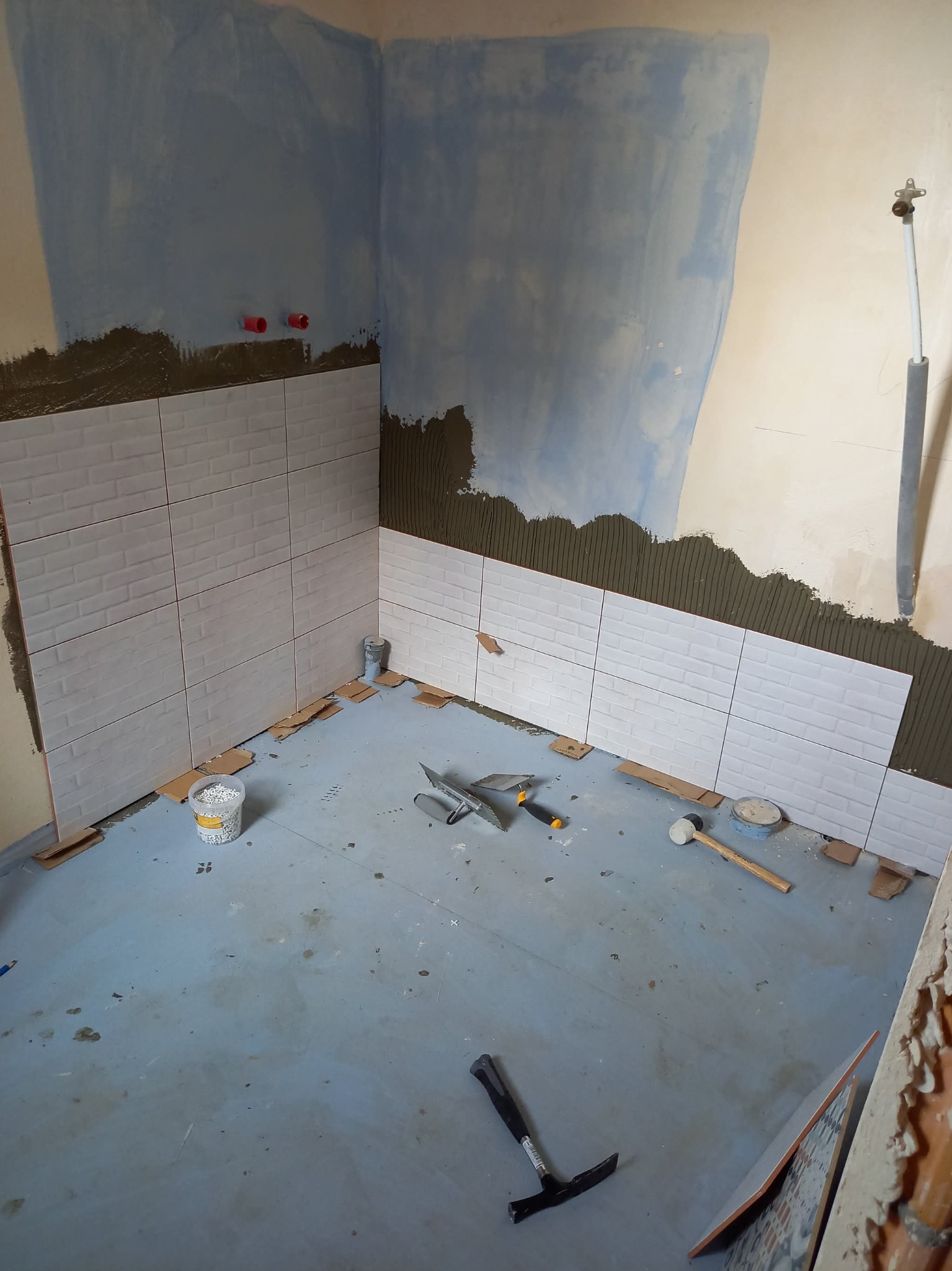 Remont łazienki w trakcie: białe płytki imitujące cegłę na ścianie, narzędzia i wiadro z krzyżykami dystansowymi na podłodze. Widoczne ślady po kleju i przygotowanie podłoża.