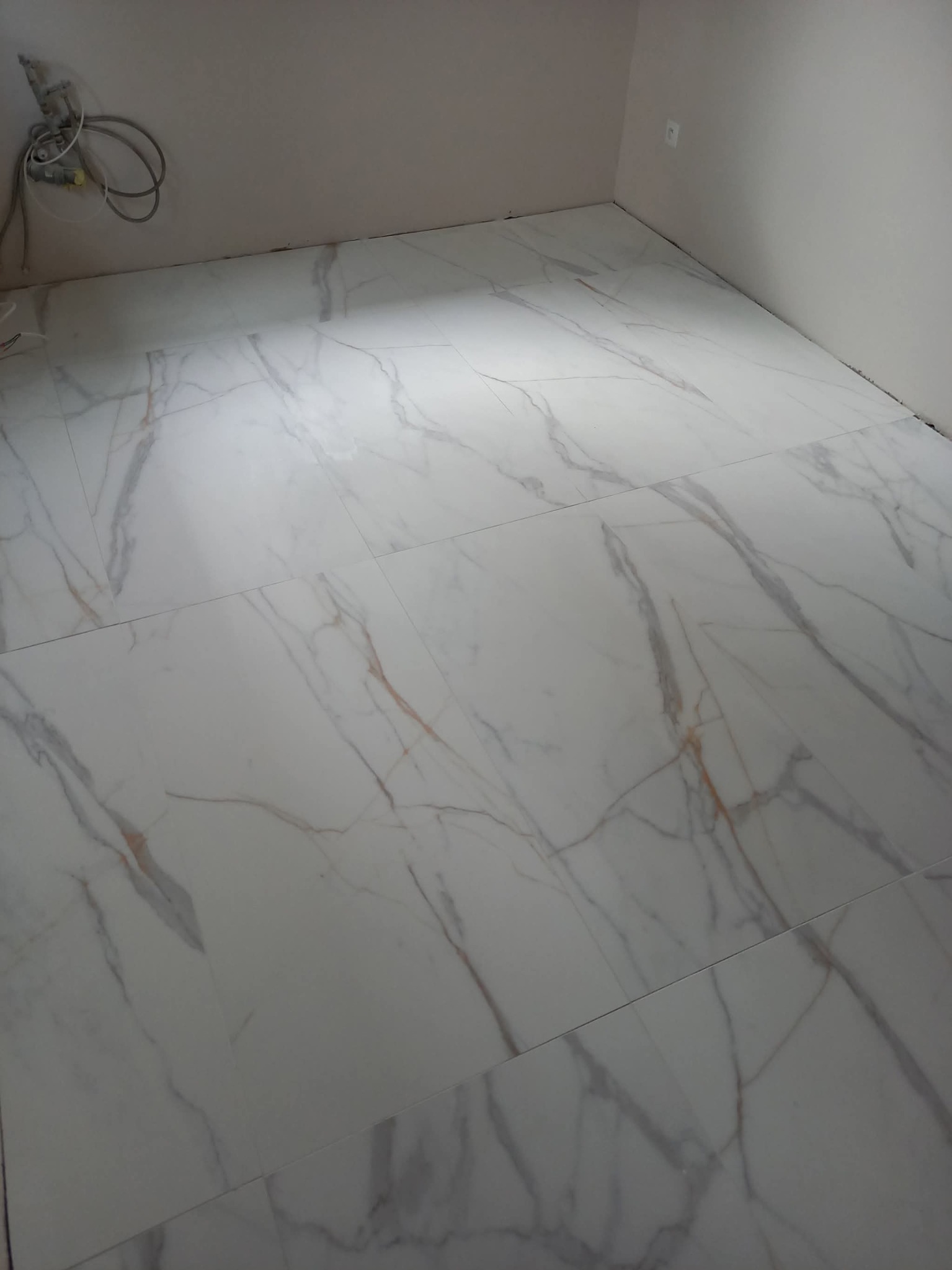 Ułożone, białe płytki podłogowe z marmurkowym wzorem w nowym pomieszczeniu. Widoczne kable wystające ze ściany. Minimalistyczny design.