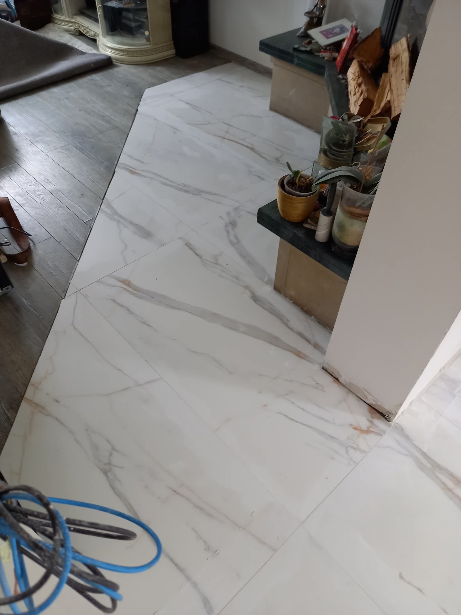 Białe płytki podłogowe z marmurowym wzorem, ułożone w pomieszczeniu z elementami dekoracyjnymi i fragmentem drewnianej podłogi. Widoczne kable w rogu kadru.
