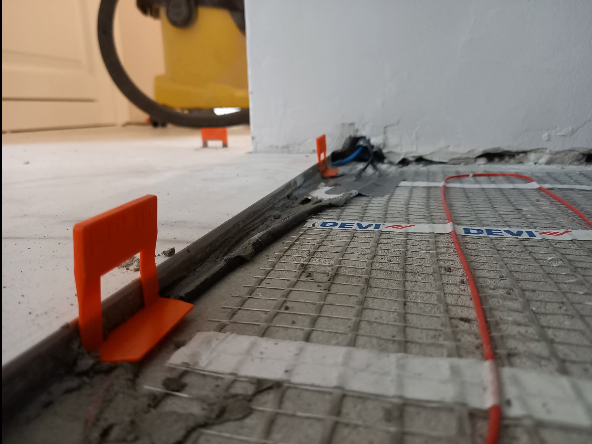 Montaż ogrzewania podłogowego z użyciem listew i klipsów poziomujących. Widoczna siatka i przewody grzewcze marki Devi. W tle odkurzacz przemysłowy.