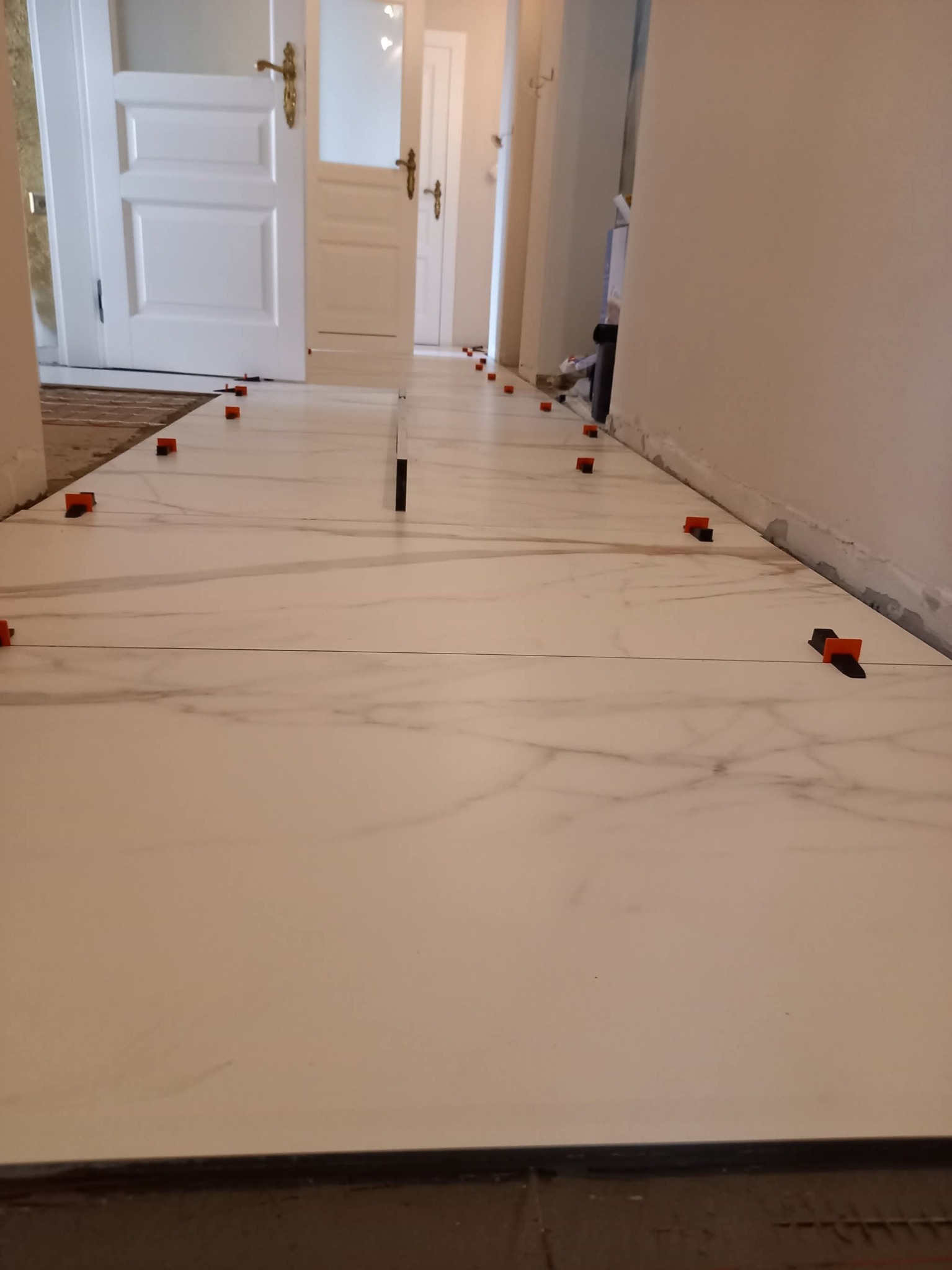 Układanie białych płytek podłogowych z marmurowym wzorem w korytarzu, z użyciem pomarańczowych klipsów poziomujących. Widoczne drzwi w tle.