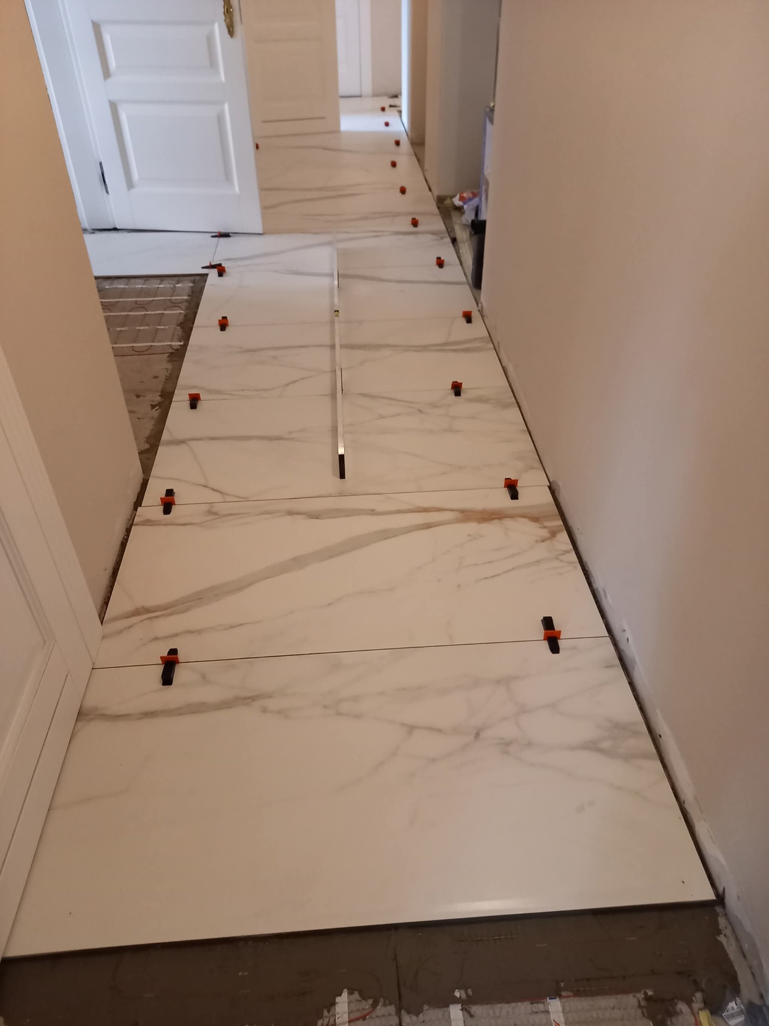 Układanie białych płytek imitujących marmur w korytarzu, z widocznymi elementami poziomowania i ogrzewaniem podłogowym. Widok z góry, perspektywa zbieżna.