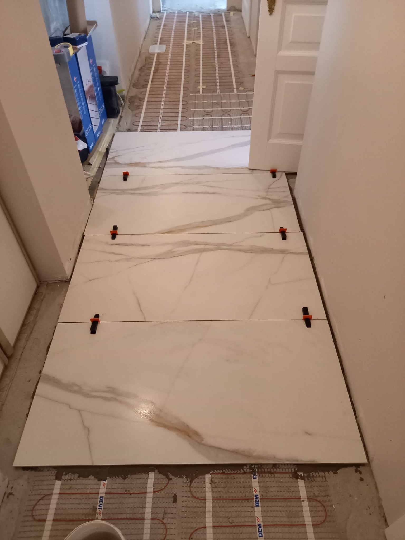 Układanie płytek podłogowych z marmurowym wzorem na ogrzewaniu podłogowym, z użyciem klipsów poziomujących. Widok z góry, wnętrze w trakcie prac wykończeniowych.