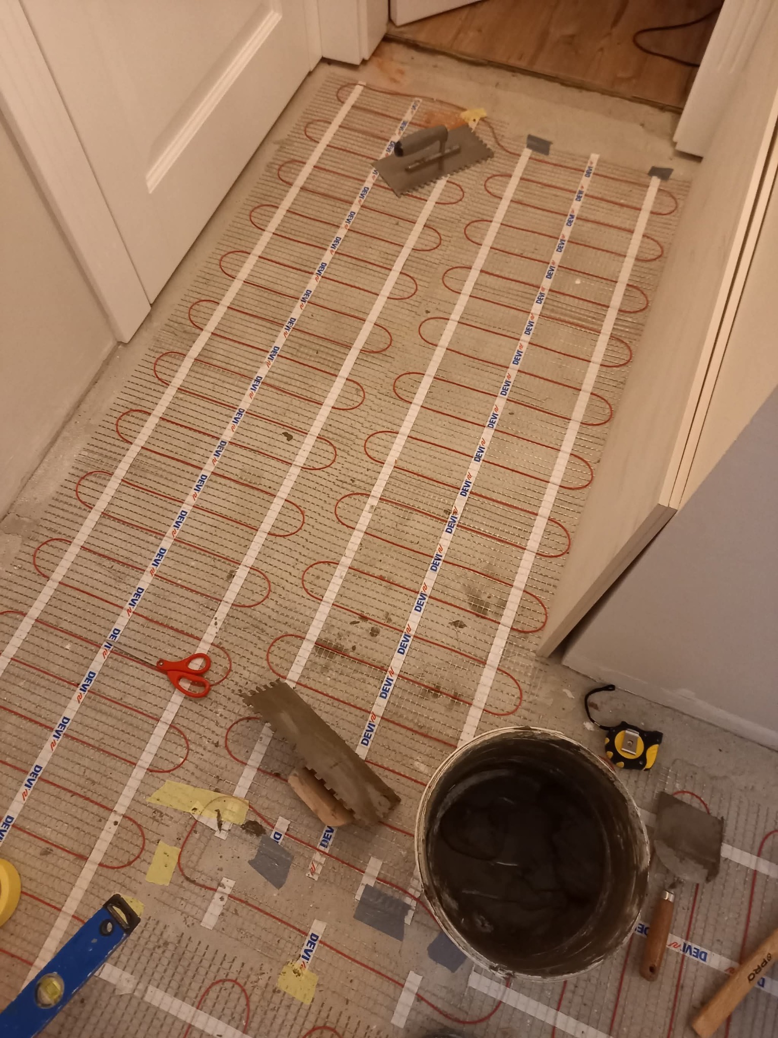 Montaż ogrzewania podłogowego: mata grzewcza z narzędziami (poziomica, paca, wiadro z zaprawą) na podłożu. Widoczne przewody grzewcze i elementy mocujące.