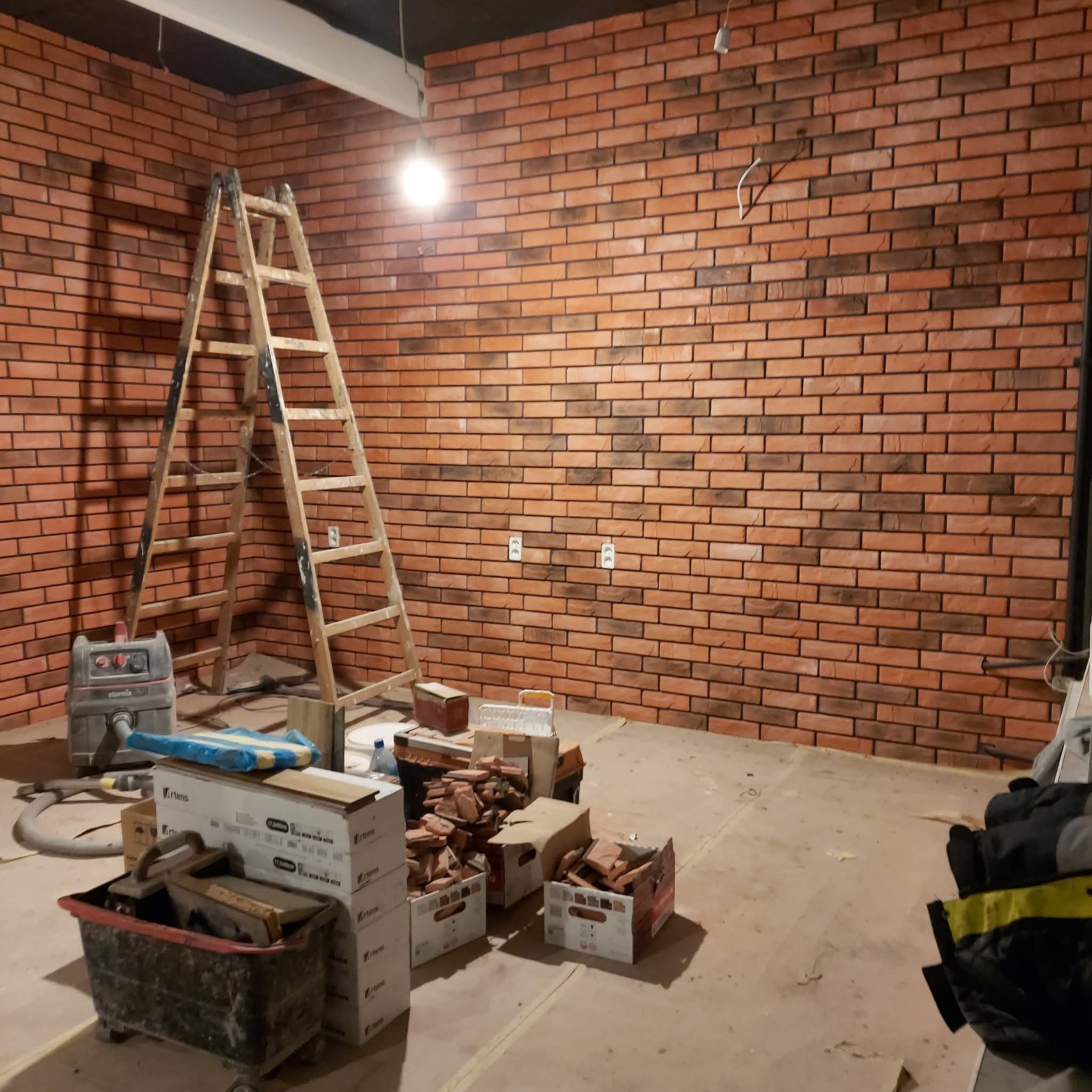 Wnętrze z ceglaną ścianą w trakcie wykańczania. Widoczna drabina, narzędzia i materiały budowlane. Oświetlenie górne, surowy stan podłogi.