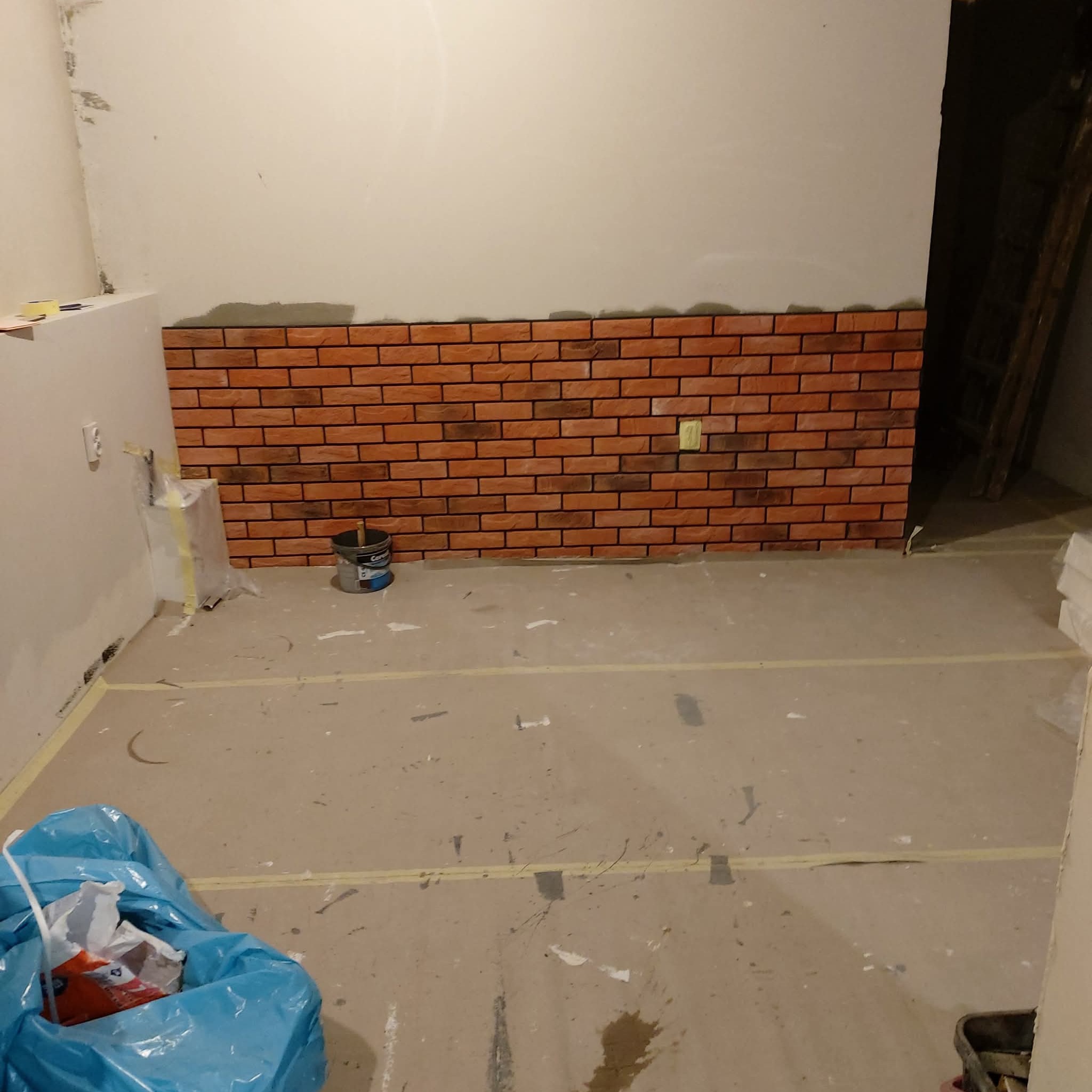 Fragment ściany z dekoracyjną okładziną imitującą czerwoną cegłę w surowym wnętrzu. Widoczne wiadro z farbą i zabezpieczenia podłogi taśmą malarską.