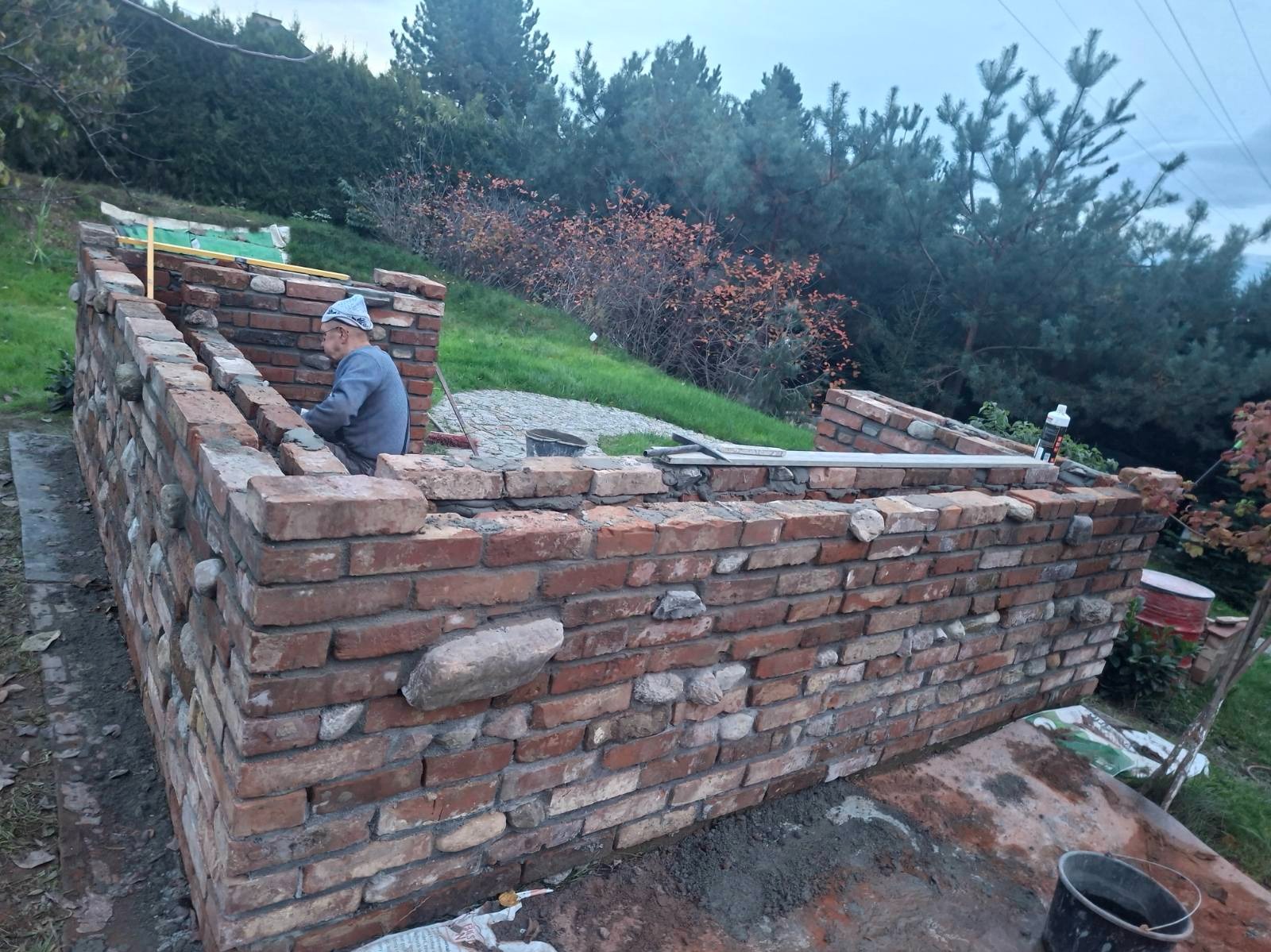 Budowa muru z czerwonej cegły i kamieni polnych na zewnątrz. Widoczny murarz w trakcie pracy, narzędzia i otoczenie z zielenią. Nietypowe połączenie materiałów.