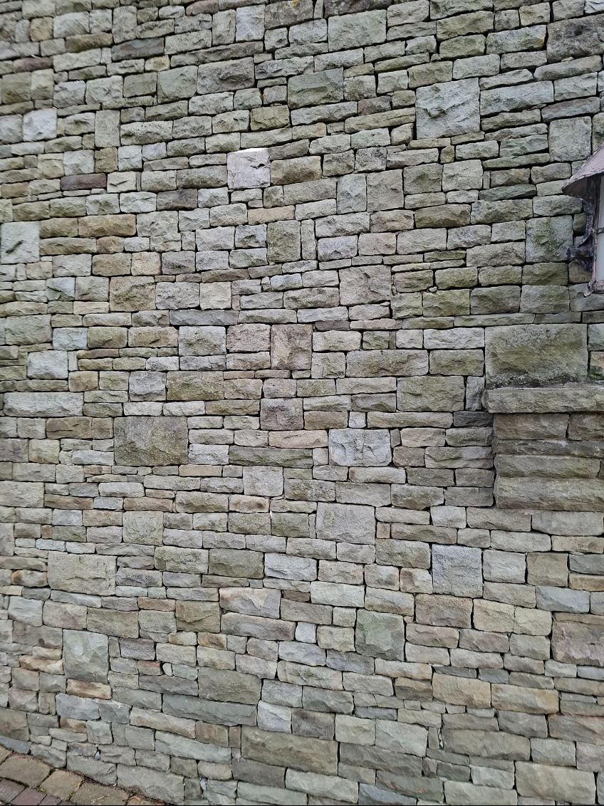 Ściana z kamienia naturalnego o nieregularnym kształcie, z widoczną lampą na elewacji. Jasne, naturalne odcienie kamienia tworzą rustykalny wygląd.