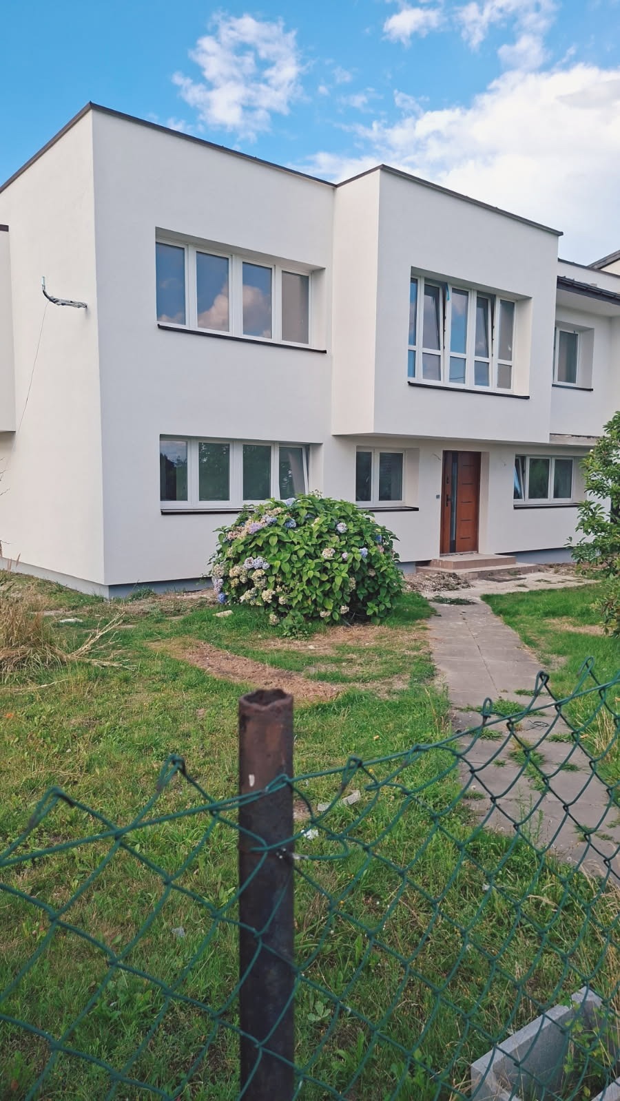 Biały, dwukondygnacyjny dom po remoncie elewacji, z nowymi oknami i odnowionym wejściem, widziany zza zielonego ogrodzenia. Hortensja przed wejściem.