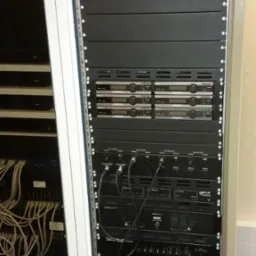Profesjonalna szafa rack z zainstalowanym sprzętem audio-wideo, widoczne kable i urządzenia różnych producentów, gotowa do pracy.