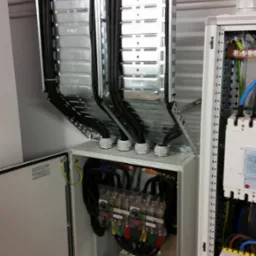 Rozdzielnia elektryczna z otwartymi drzwiami, widoczne kable w metalowych korytkach i zabezpieczenia. Profesjonalny montaż instalacji elektrycznej w budynku.