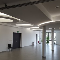 NEXA ENERGY Leszek Korycki - Nowoczesne oświetlenie sufitowe w przestronnym, jasnym wnętrzu. Okrągłe lampy wkomponowane w podwieszany sufit, kolumny i duże okna w tle.