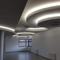 NEXA ENERGY Leszek Korycki - Nowoczesne oświetlenie sufitowe z okrągłymi lampami LED w przestronnym, jasnym wnętrzu. Widoczne okna z żaluzjami i kolumna podtrzymująca sufit.