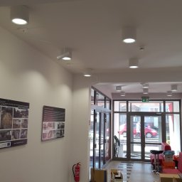 NEXA ENERGY Leszek Korycki - Białe, okrągłe lampy sufitowe w jasnym holu budynku, z widocznymi plakatami na ścianie i kamerą monitoringu. Nowoczesne oświetlenie w przestrzeni publicznej.