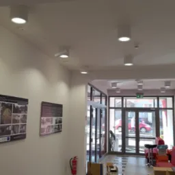 Białe, okrągłe lampy sufitowe w jasnym holu budynku, z widocznymi plakatami na ścianie i kamerą monitoringu. Nowoczesne oświetlenie w przestrzeni publicznej.