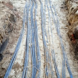 NEXA ENERGY Leszek Korycki - Kable w wykopie, zabezpieczone niebieskimi rurami karbowanymi. Widoczne ślady ziemi na kablach. Wykop z ceglanym murem w tle.