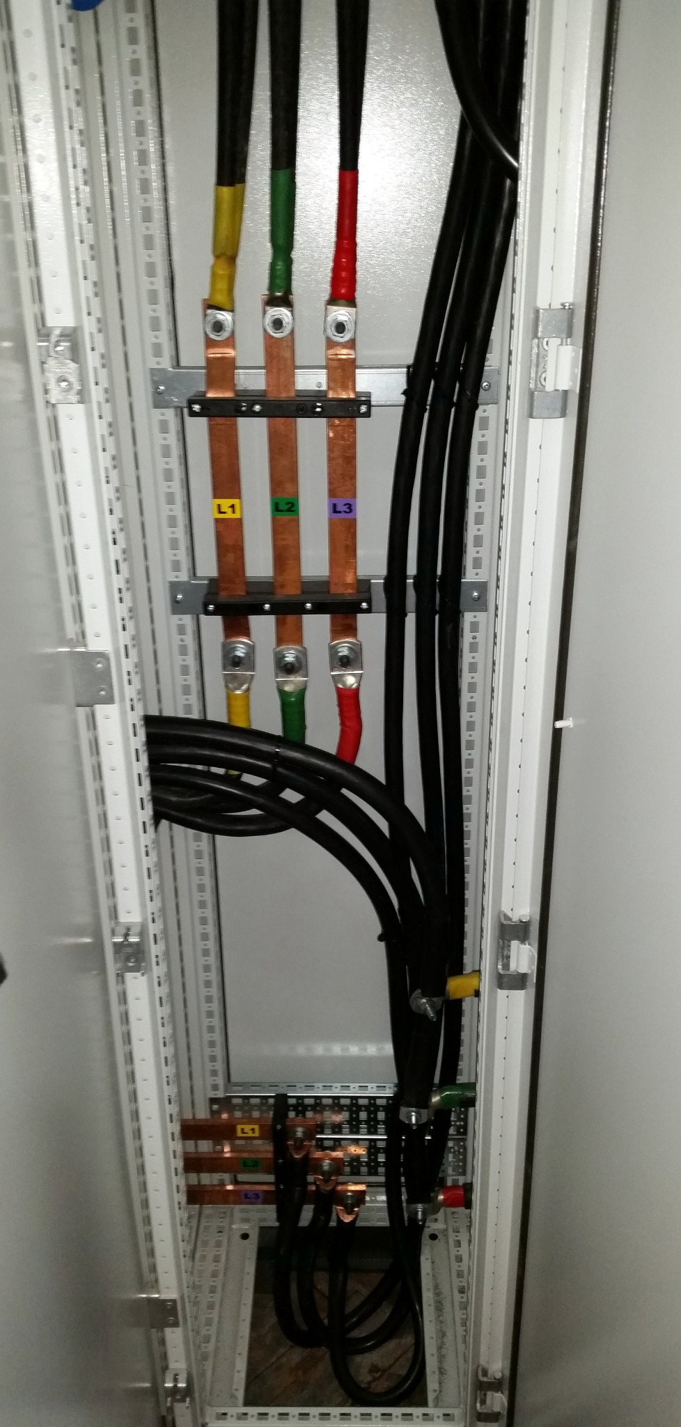 Rozdzielnica elektryczna wewnątrz, z przewodami oznaczonymi kolorami (żółty, zielony, czerwony) i etykietami L1, L2, L3. Widoczne szyny miedziane i czarne kable.