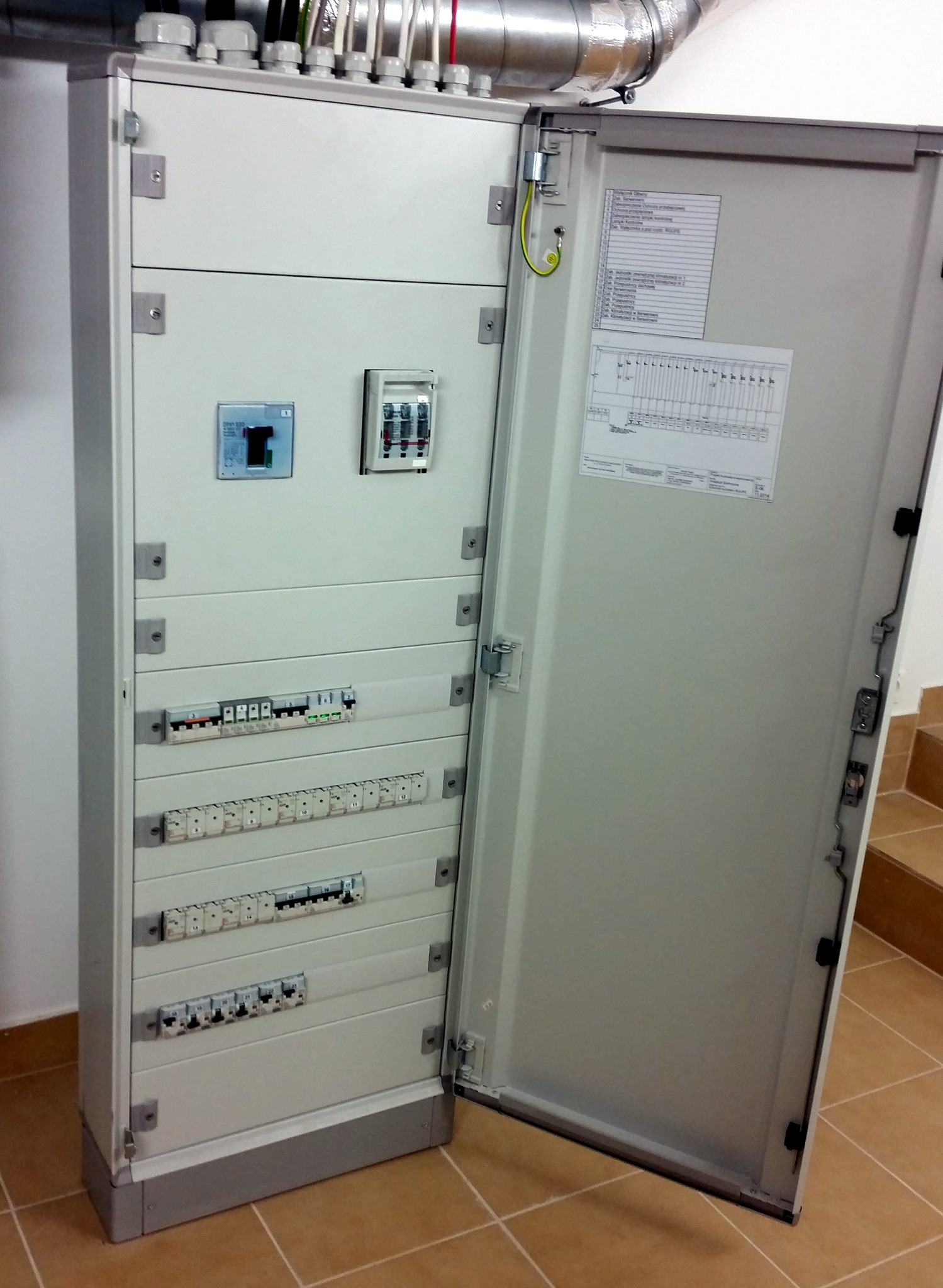 Otwarta, szara szafa elektryczna z bezpiecznikami i wyłącznikami, widoczna instalacja w rurze na suficie, płytki na podłodze, schody w tle.