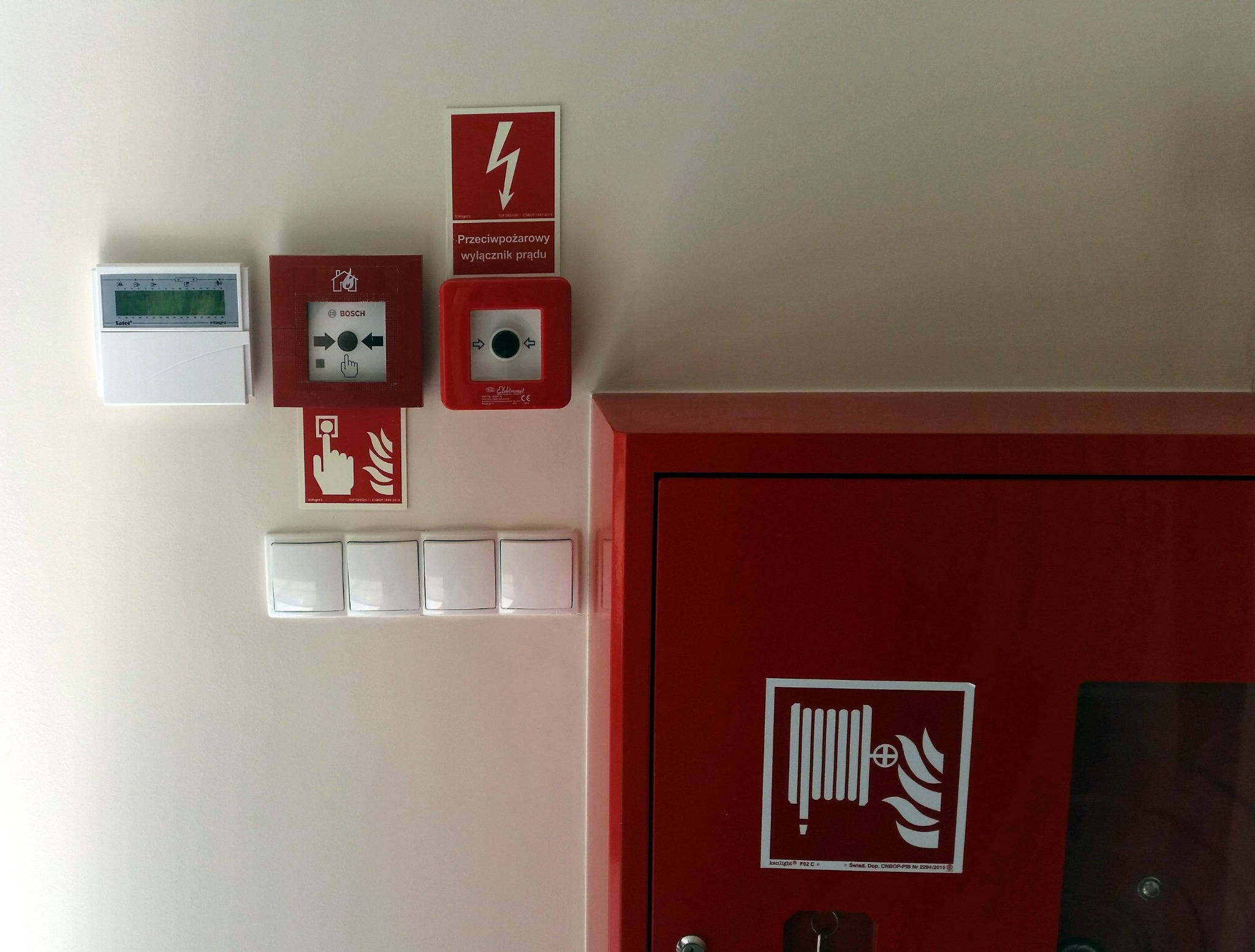 Elementy systemu przeciwpożarowego: centrala alarmowa, przycisk alarmowy Bosch, wyłącznik prądu i czerwona szafka hydrantowa z instrukcją użycia na białej ścianie.