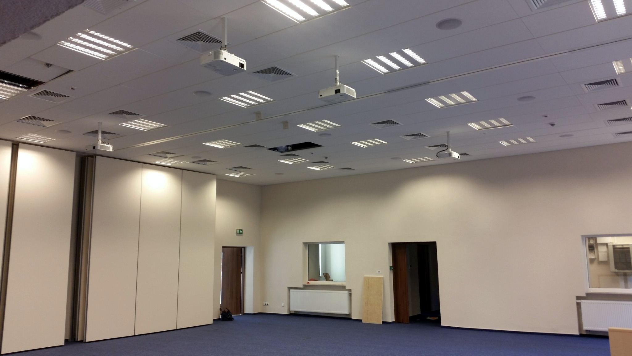 Sufit biura z równomiernie rozmieszczonymi lampami i projektorami, niebieska wykładzina podłogowa, białe ściany i składane ścianki działowe.