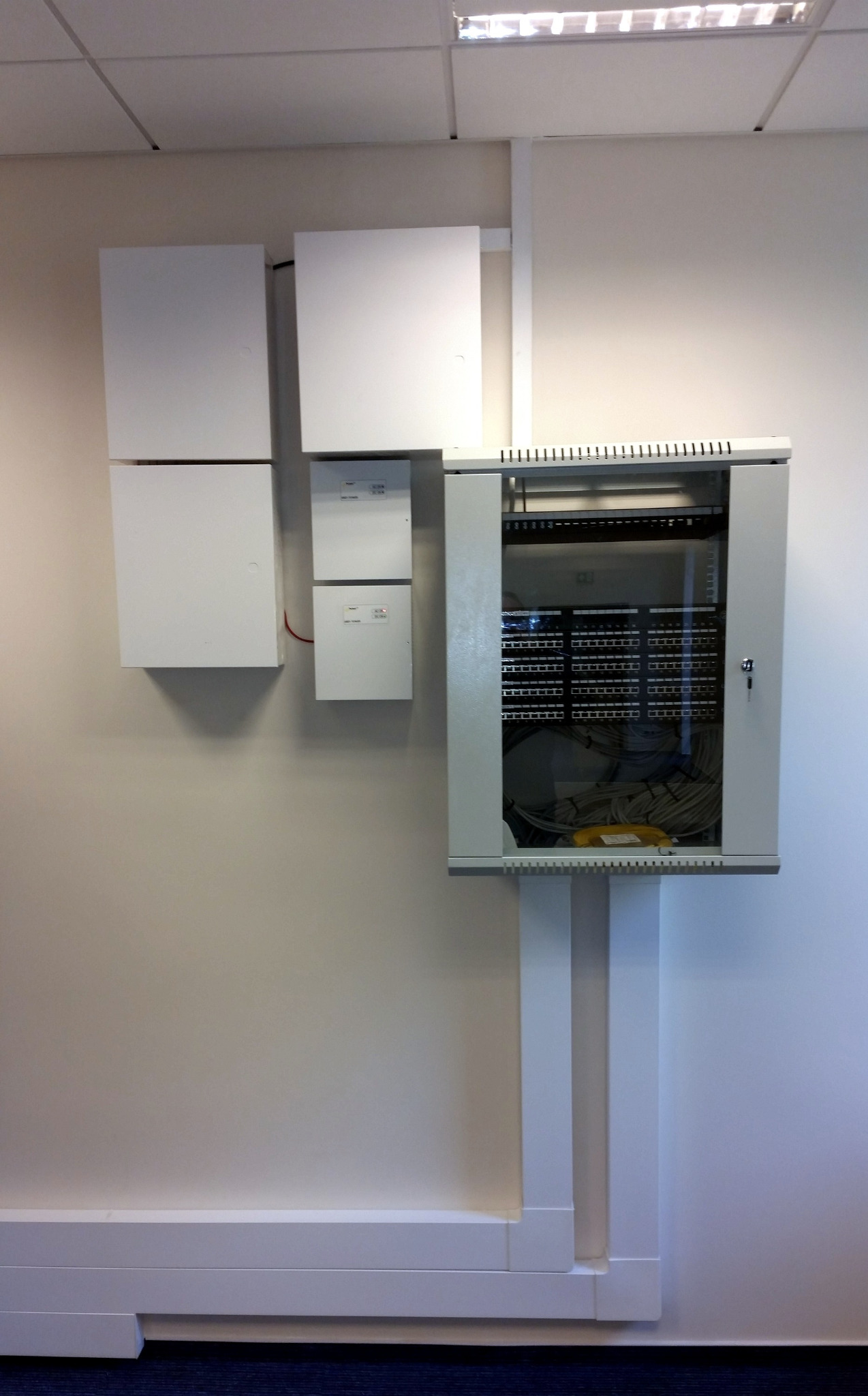 Zamknięta szafa rack z widocznymi kablami sieciowymi zamontowana na ścianie, obok skrzynki elektryczne. Białe ściany, listwy przypodłogowe i niebieska wykładzina podłogowa.
