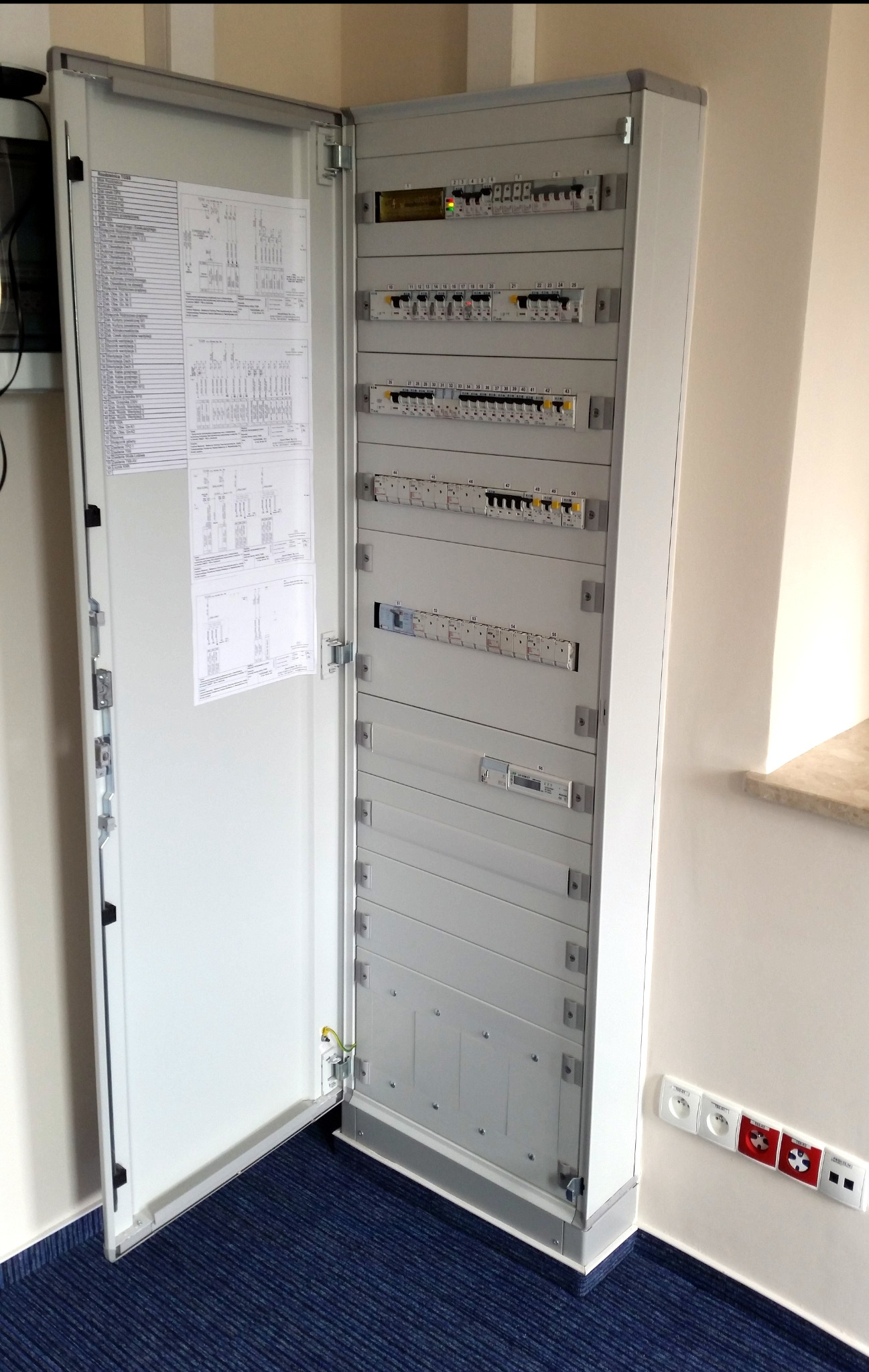 Otwarta, szara szafa elektryczna z widocznymi bezpiecznikami i schematami, zamontowana w pomieszczeniu z niebieską wykładziną i gniazdkami elektrycznymi.