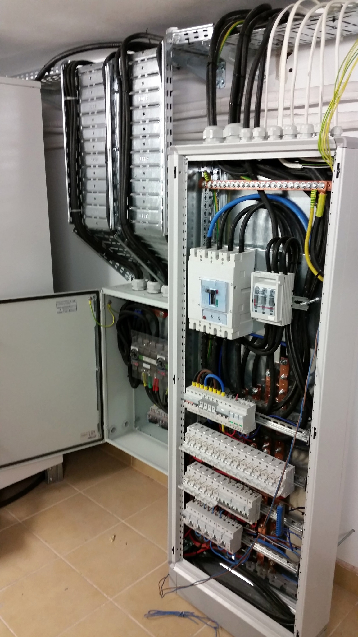 Rozdzielnica elektryczna z licznymi przewodami i zabezpieczeniami, widoczne kable prowadzone w korytach. Otwarta szafa z instalacją elektryczną w budynku.