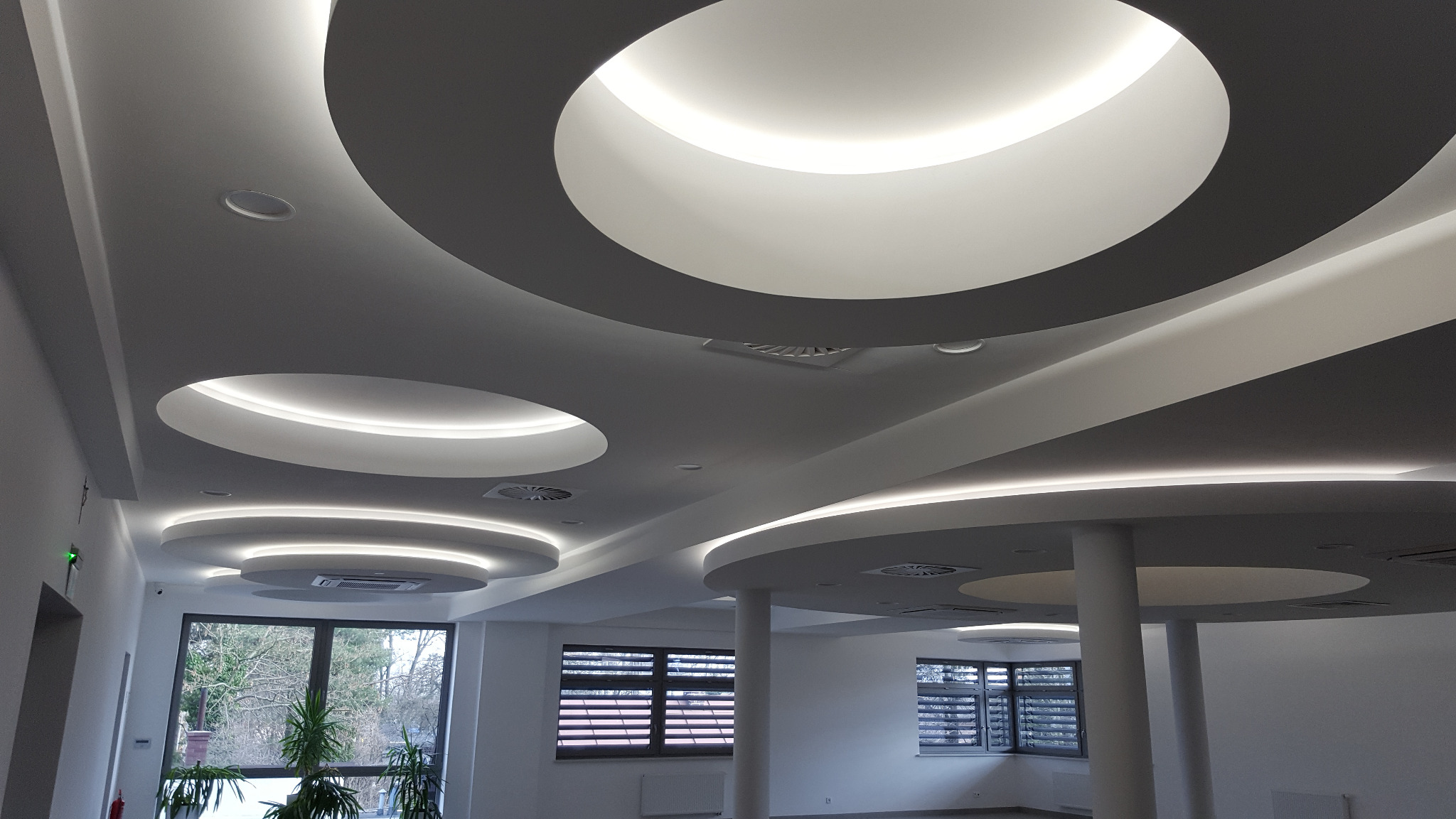 Nowoczesny sufit podwieszany z okrągłymi elementami oświetleniowymi LED, widok z dołu, jasne wnętrze z oknami i kolumnami.