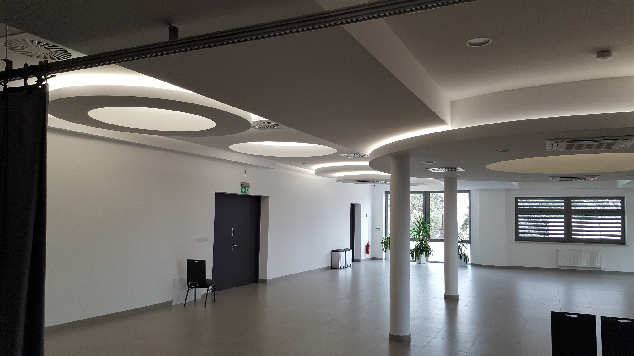 Nowoczesne oświetlenie sufitowe w przestronnym, jasnym wnętrzu. Okrągłe lampy wkomponowane w podwieszany sufit, kolumny i duże okna w tle.