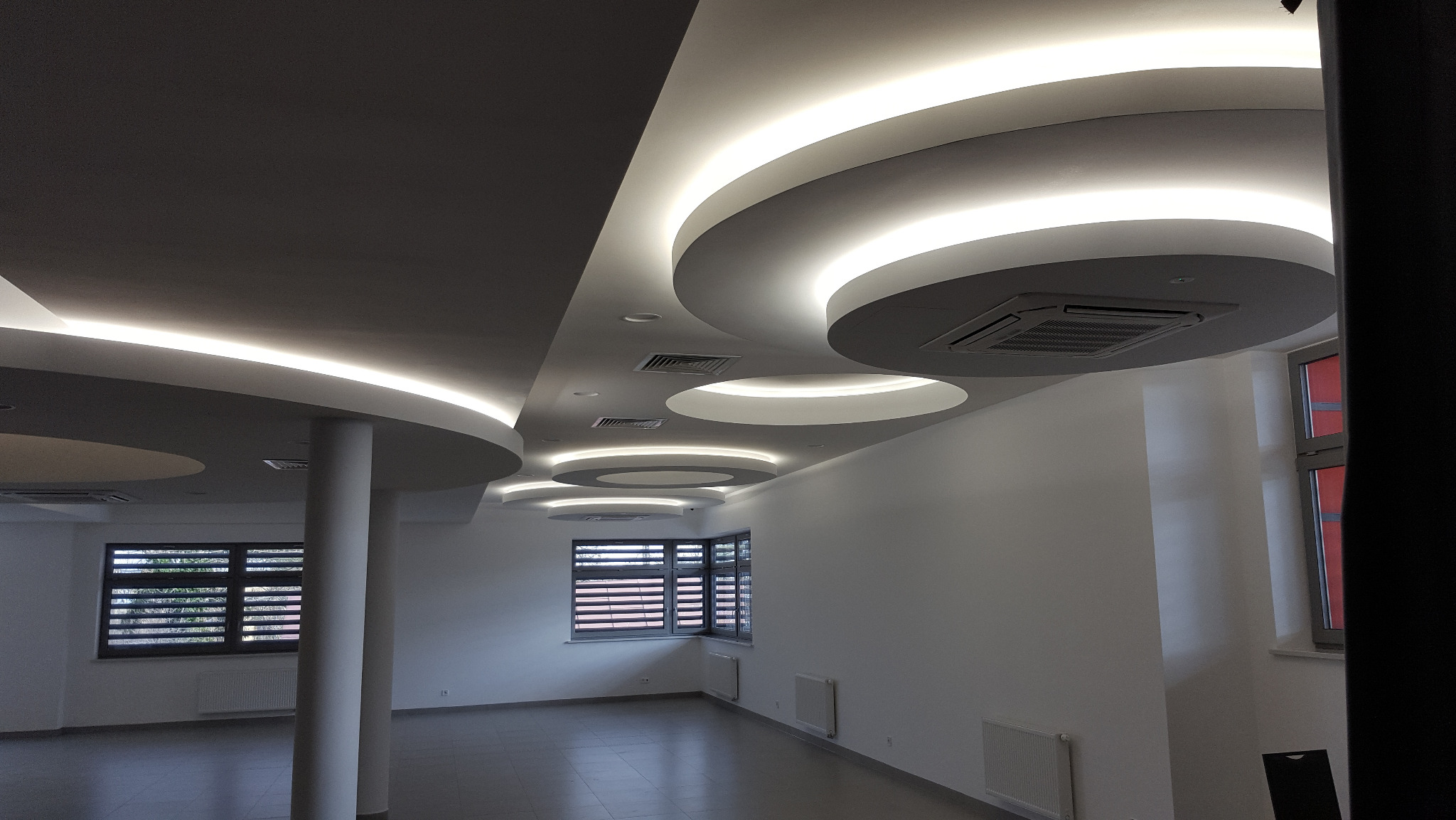 Nowoczesne oświetlenie sufitowe z okrągłymi lampami LED w przestronnym, jasnym wnętrzu. Widoczne okna z żaluzjami i kolumna podtrzymująca sufit.
